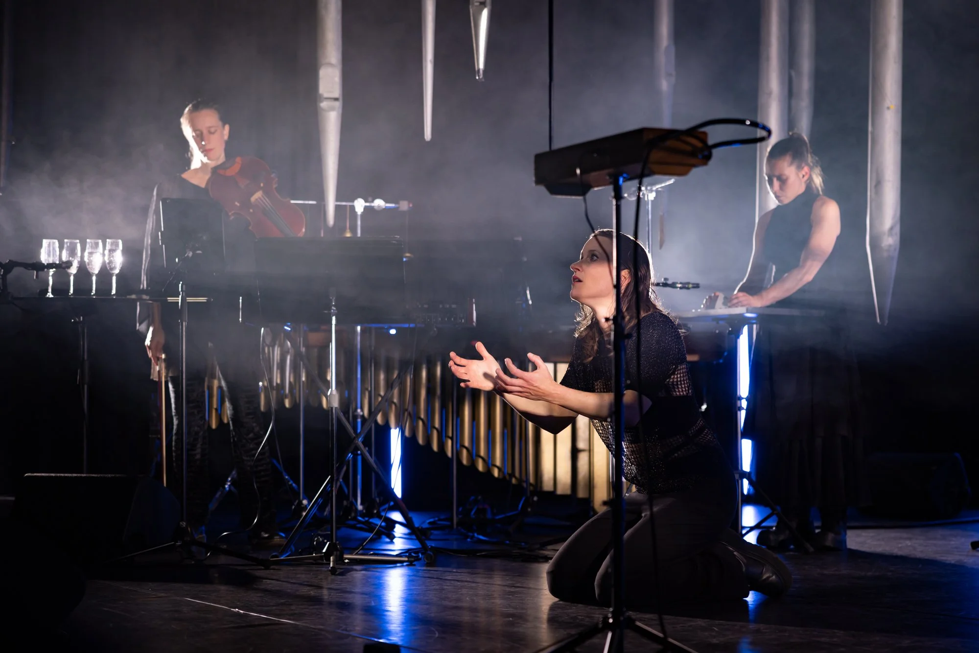 Three women performing music on stage, one kneeling with hands extended, and two playing instruments in the background amidst stage lighting and fog.