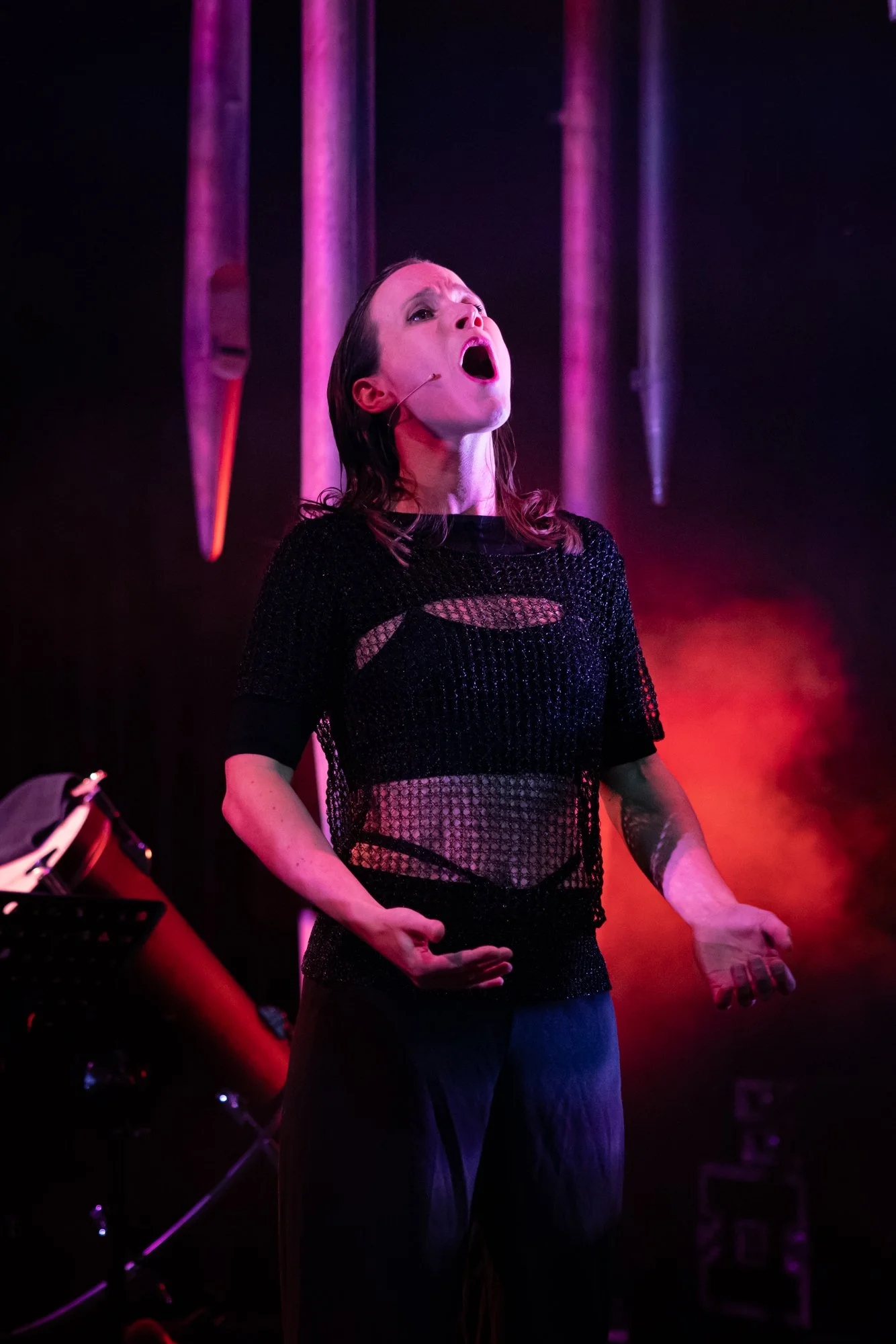 Performer singing passionately on stage, wearing a black, see-through, ripped shirt, under pink and purple stage lights.