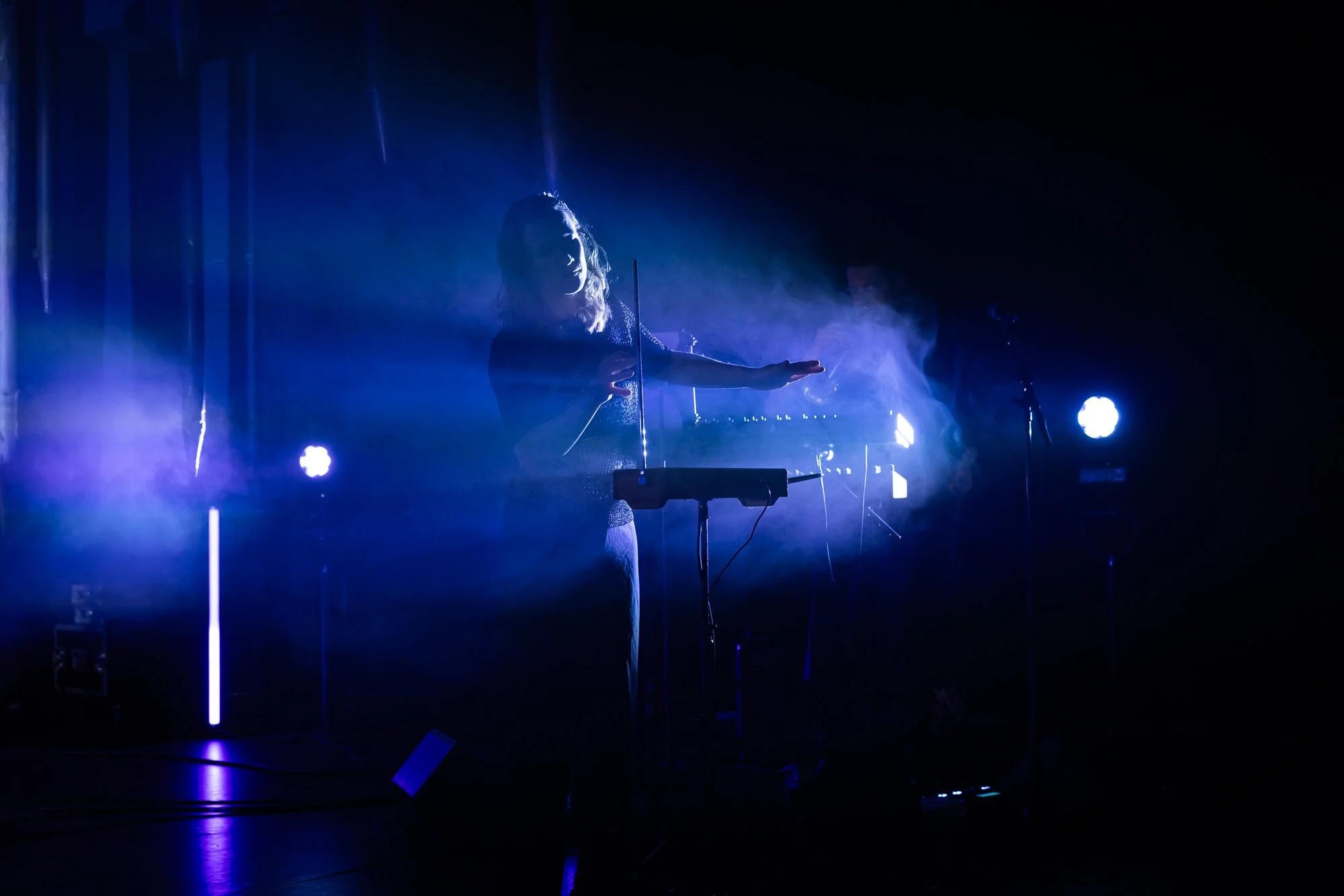 A woman performing on stage with blue and purple lighting, standing behind a keyboard and a microphone.