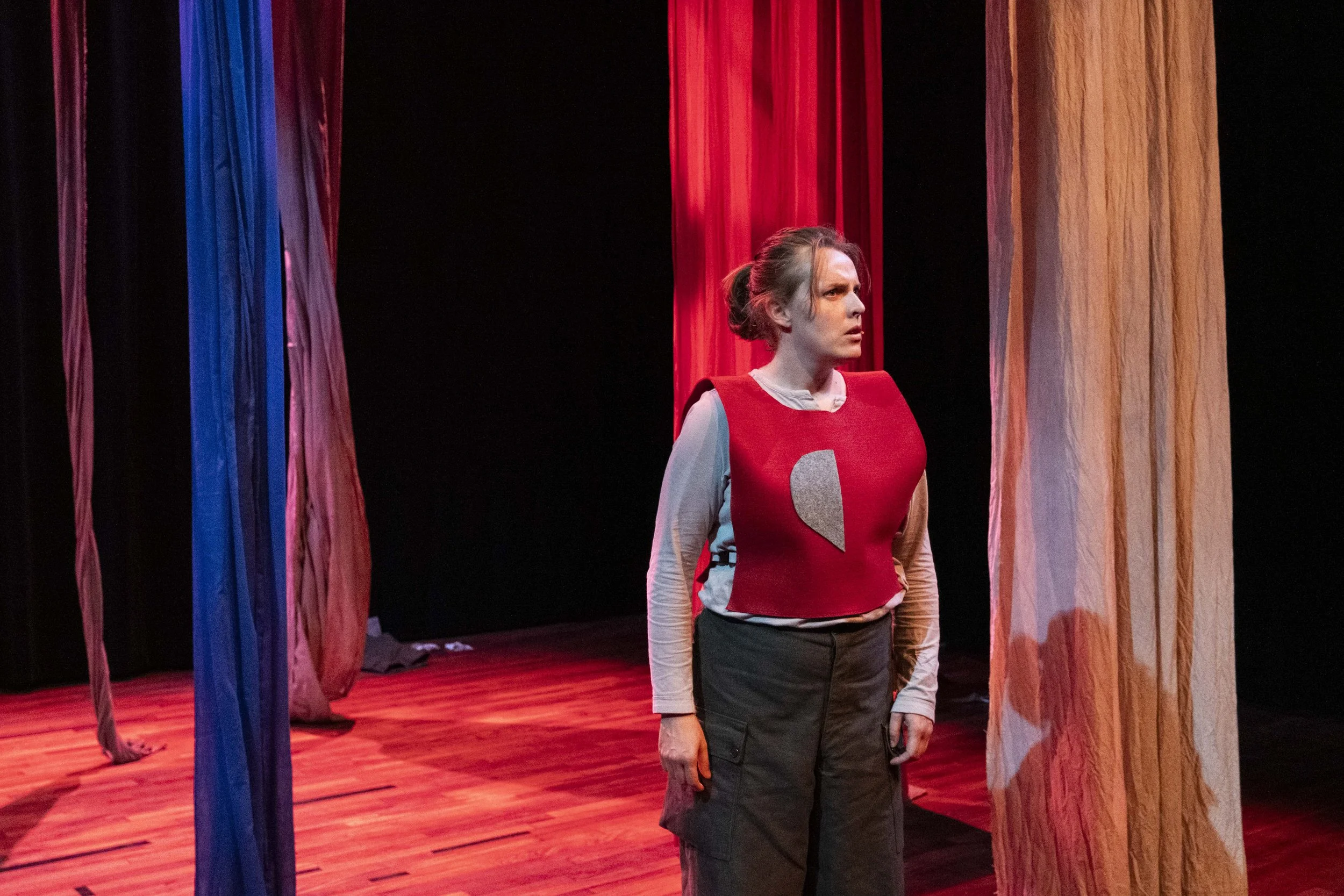 A woman standing on a stage surrounded by multicolored curtains, wearing a red vest and cargo pants, appearing to be in a theatrical performance.