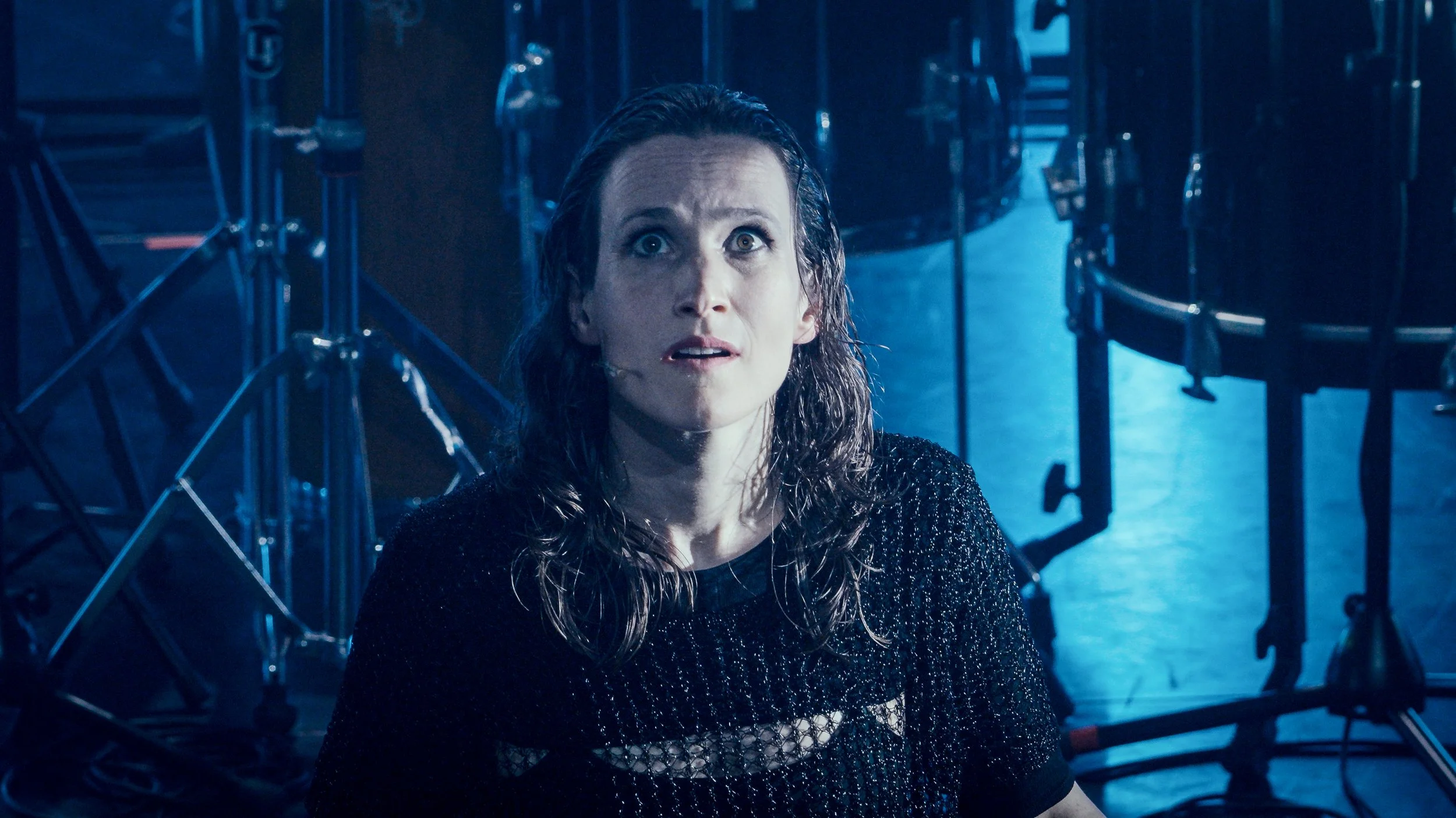A woman with wet hair looks upward with a surprised or distressed expression in a dark studio with musical equipment in the background, illuminated by blue lighting.