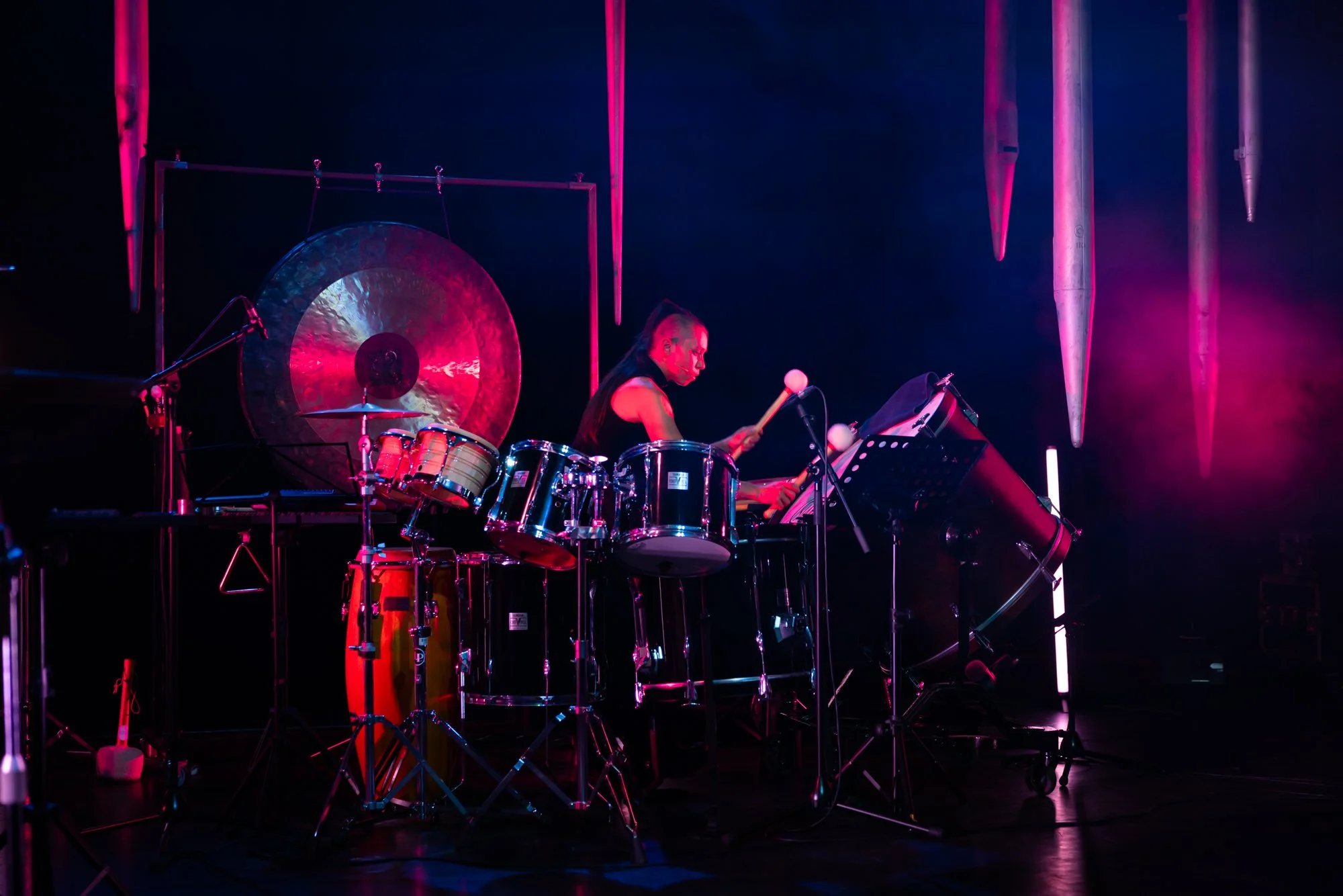 A musician playing percussion instruments on stage with pink and purple lighting, including a gong, drums, and chimes.