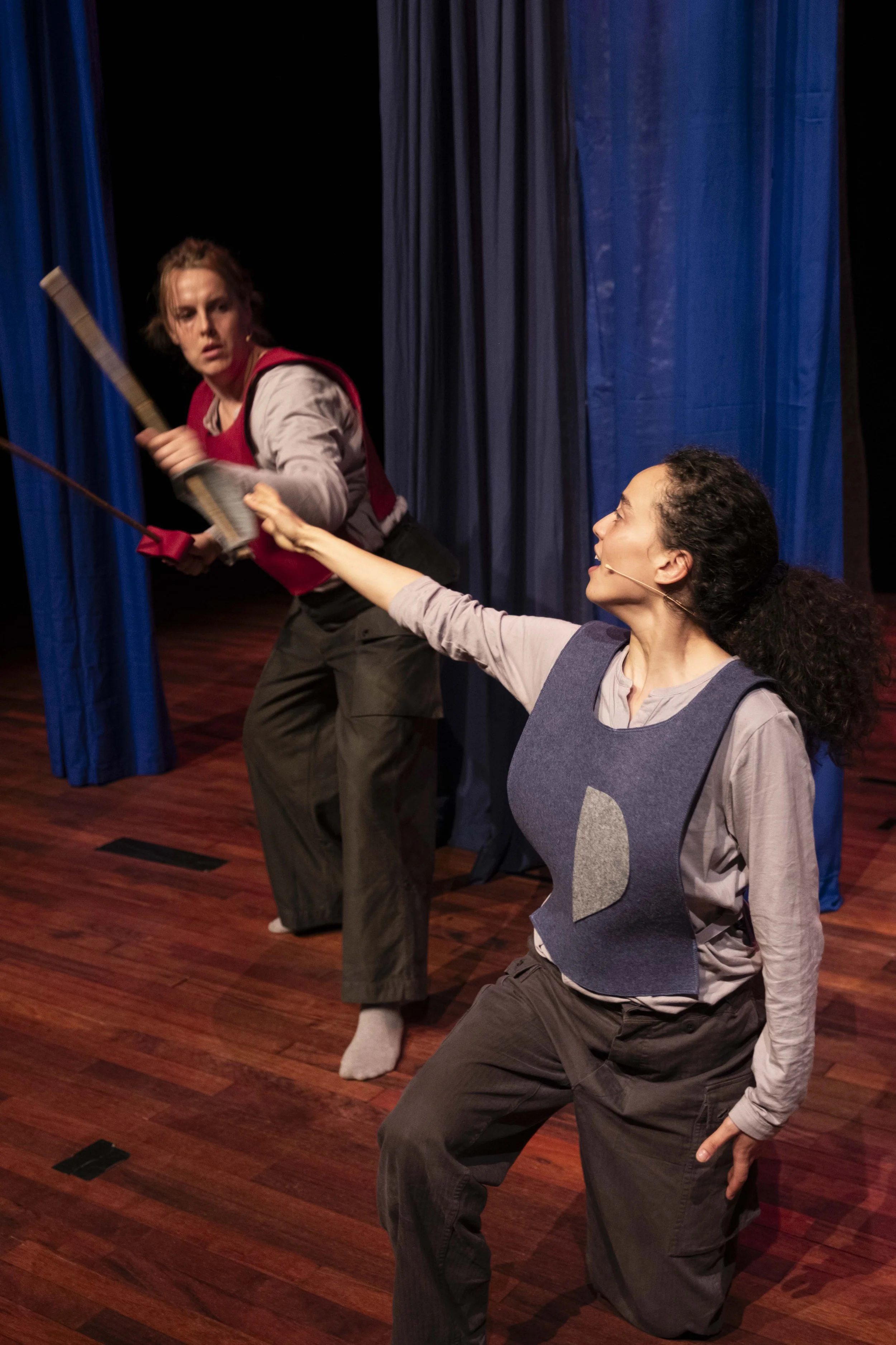 Two women engaged in a theatrical fight scene on stage, with one woman holding a weapon and the other woman reaching out.