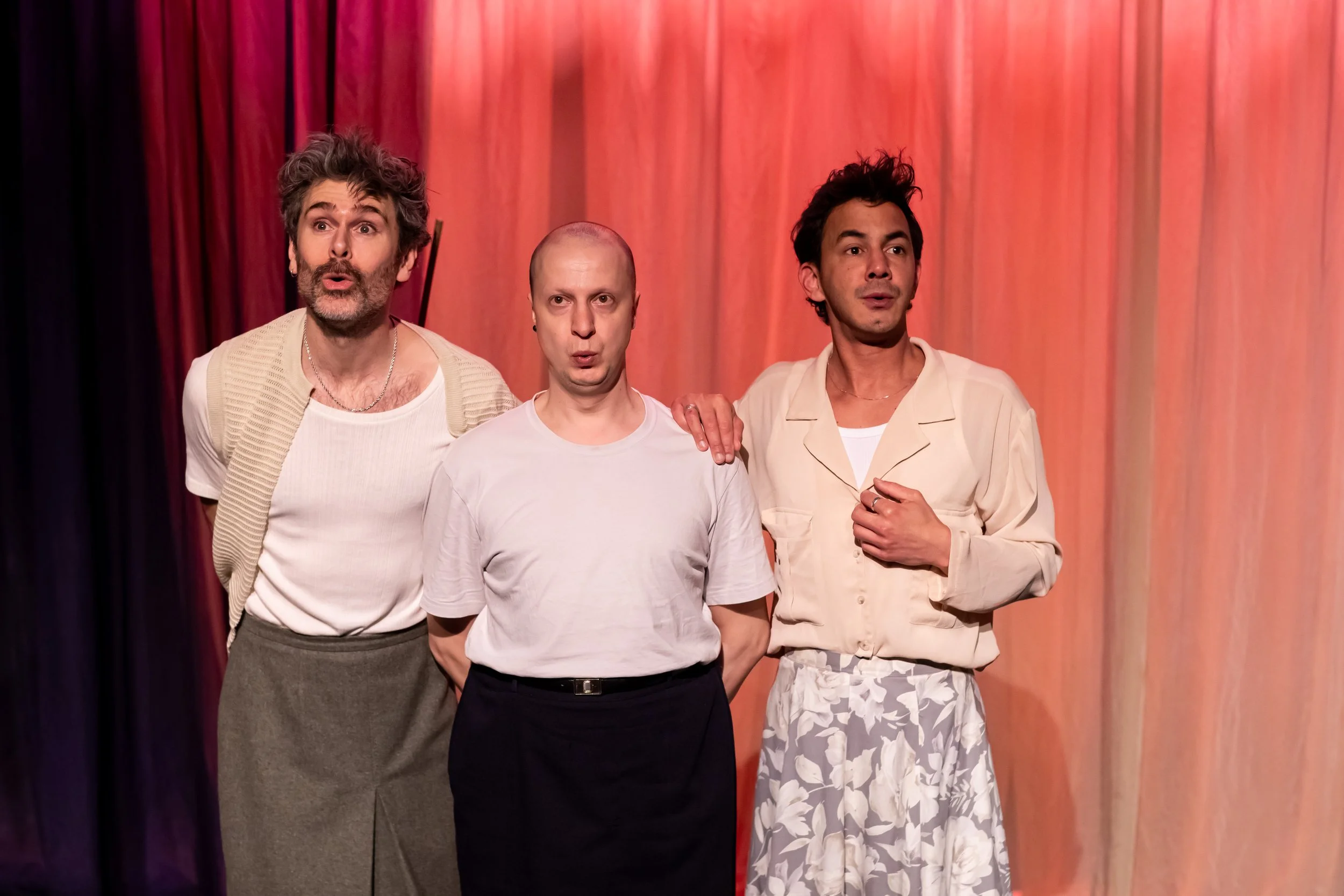 Three men standing in front of a red stage curtain, wearing casual clothing, with varied expressions and gestures.