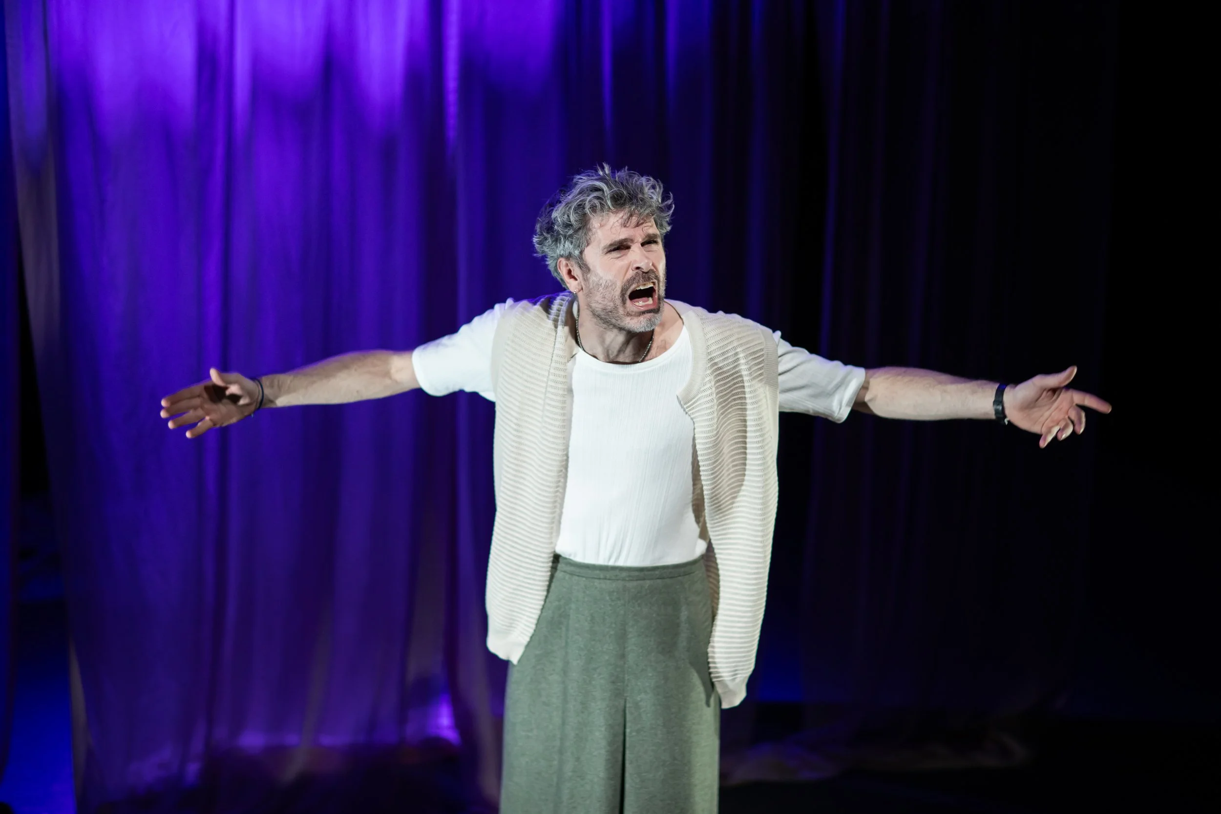 Man performing on stage with purple curtains, arms outstretched, wearing a white shirt, beige vest, and gray pants, conveying emotion.