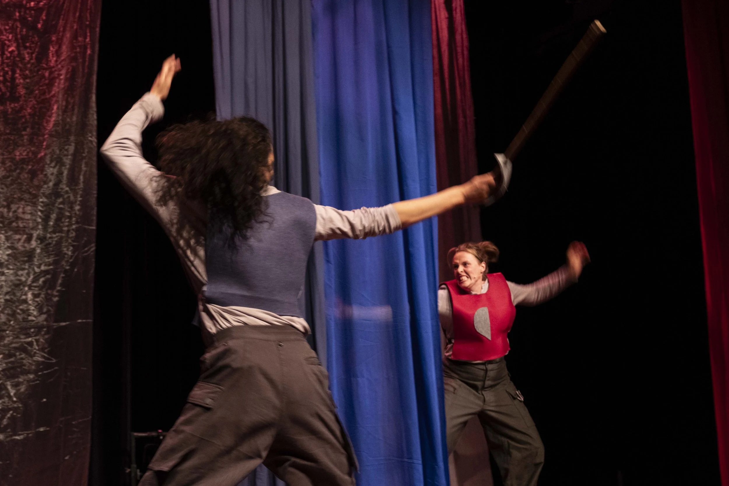 Two women in energetic poses on a stage with colored curtains, one wielding a sword and the other dressed in a red vest, engaging in a theatrical performance.