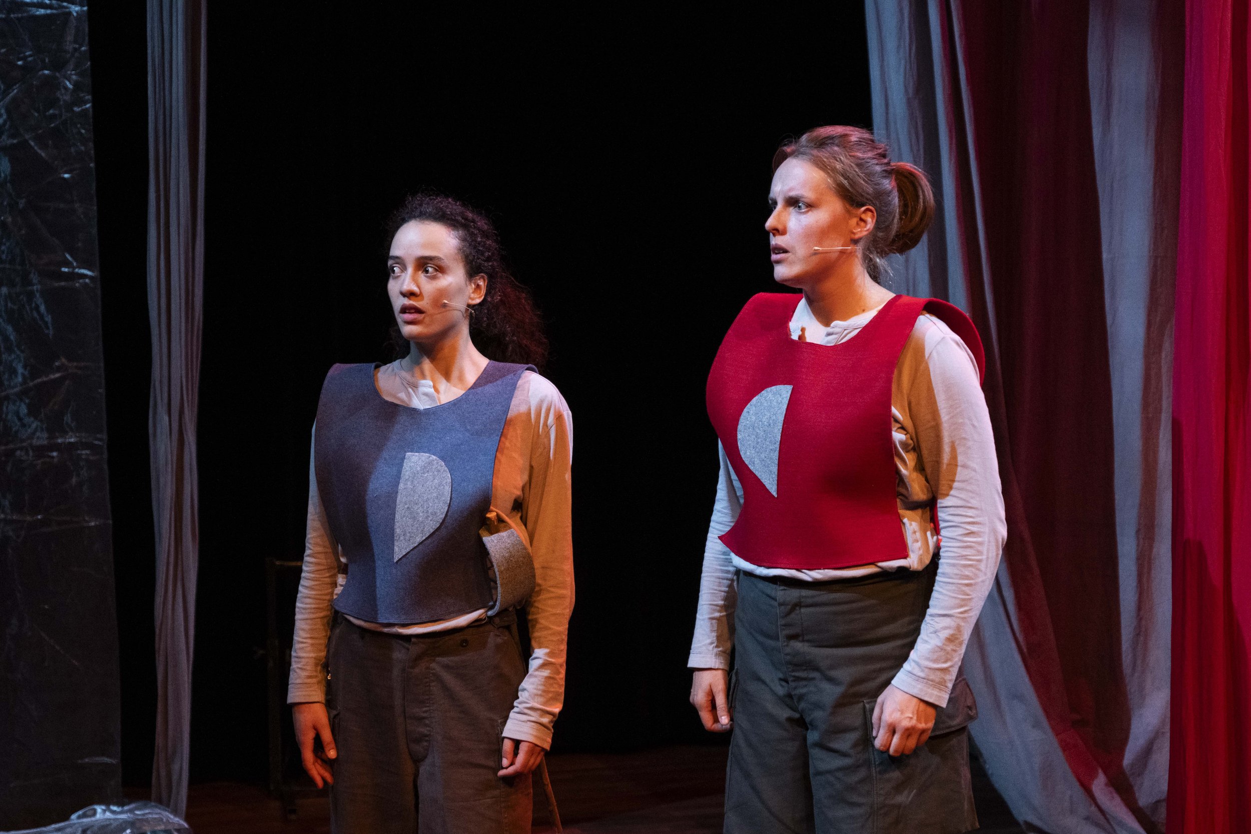 Two women on stage wearing futuristic costumes with geometric designs, appearing surprised or concerned during a theatrical performance.