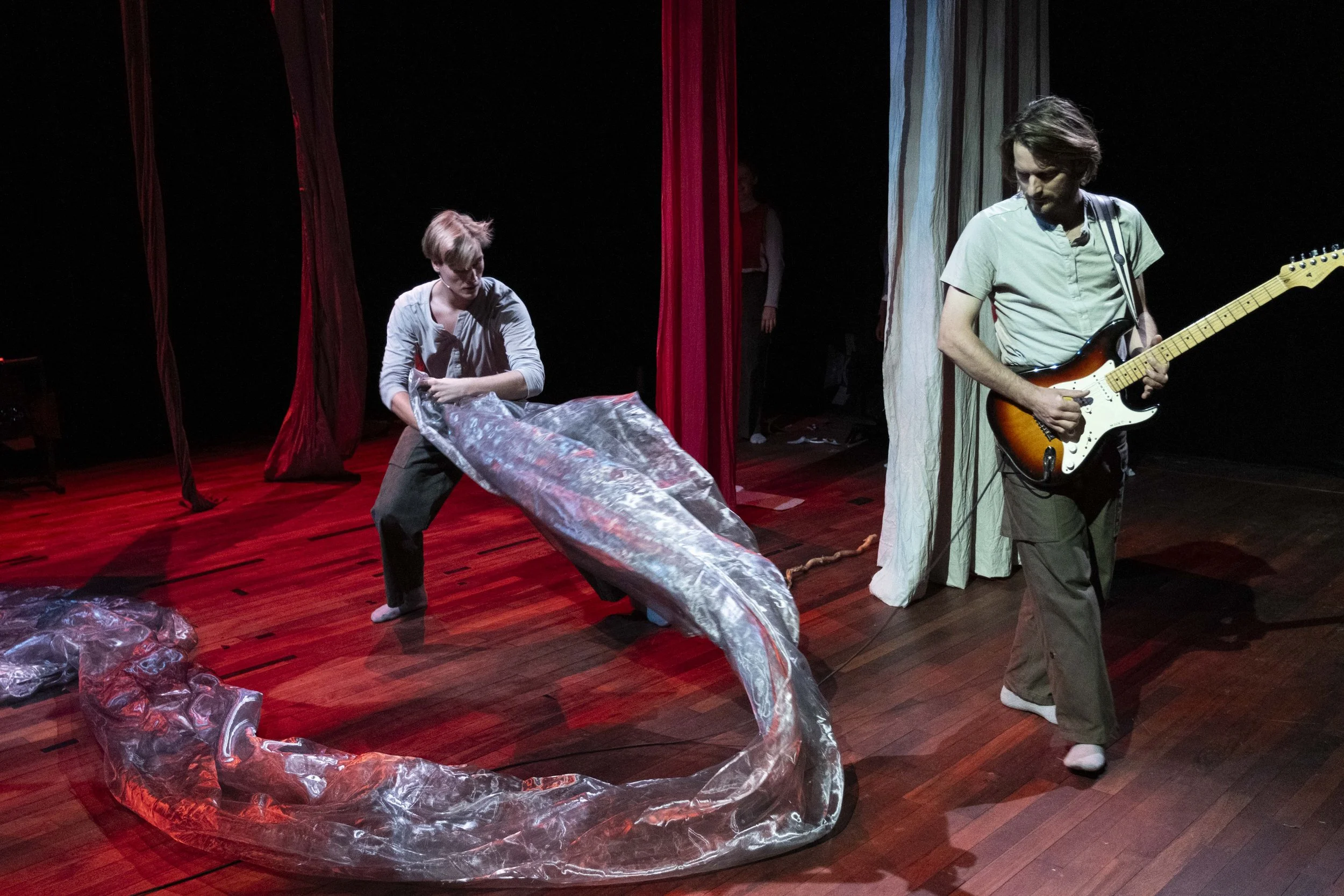 Two performers on stage, one is bending with a plastic sheet, the other playing an electric guitar, with red curtains in the background.