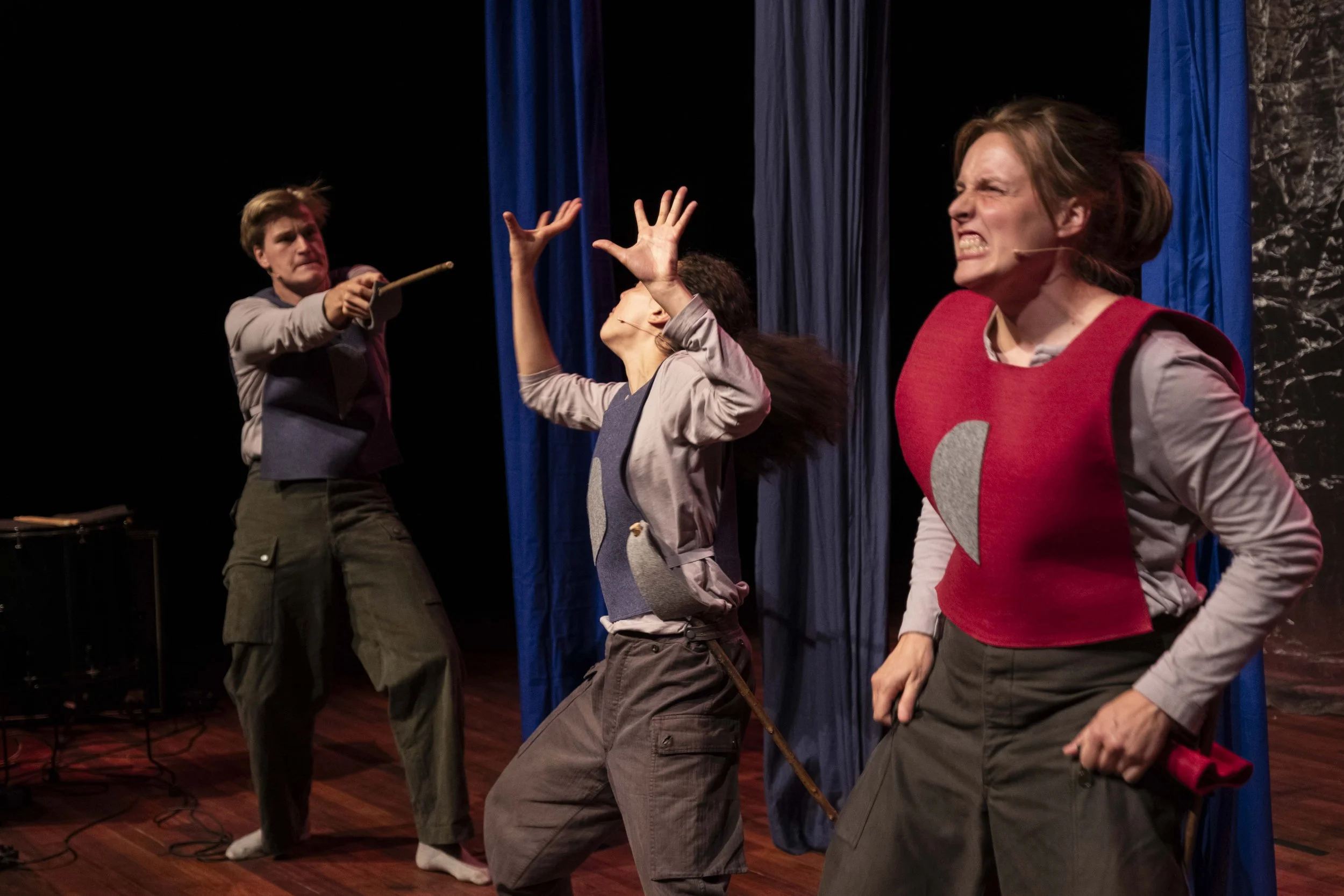 Three people performing in a theatrical play on stage: one woman is holding her hands up and leaning back, another woman in red is clenching her fists with an angry expression, and a man in the background is pointing a stick with a determined look.