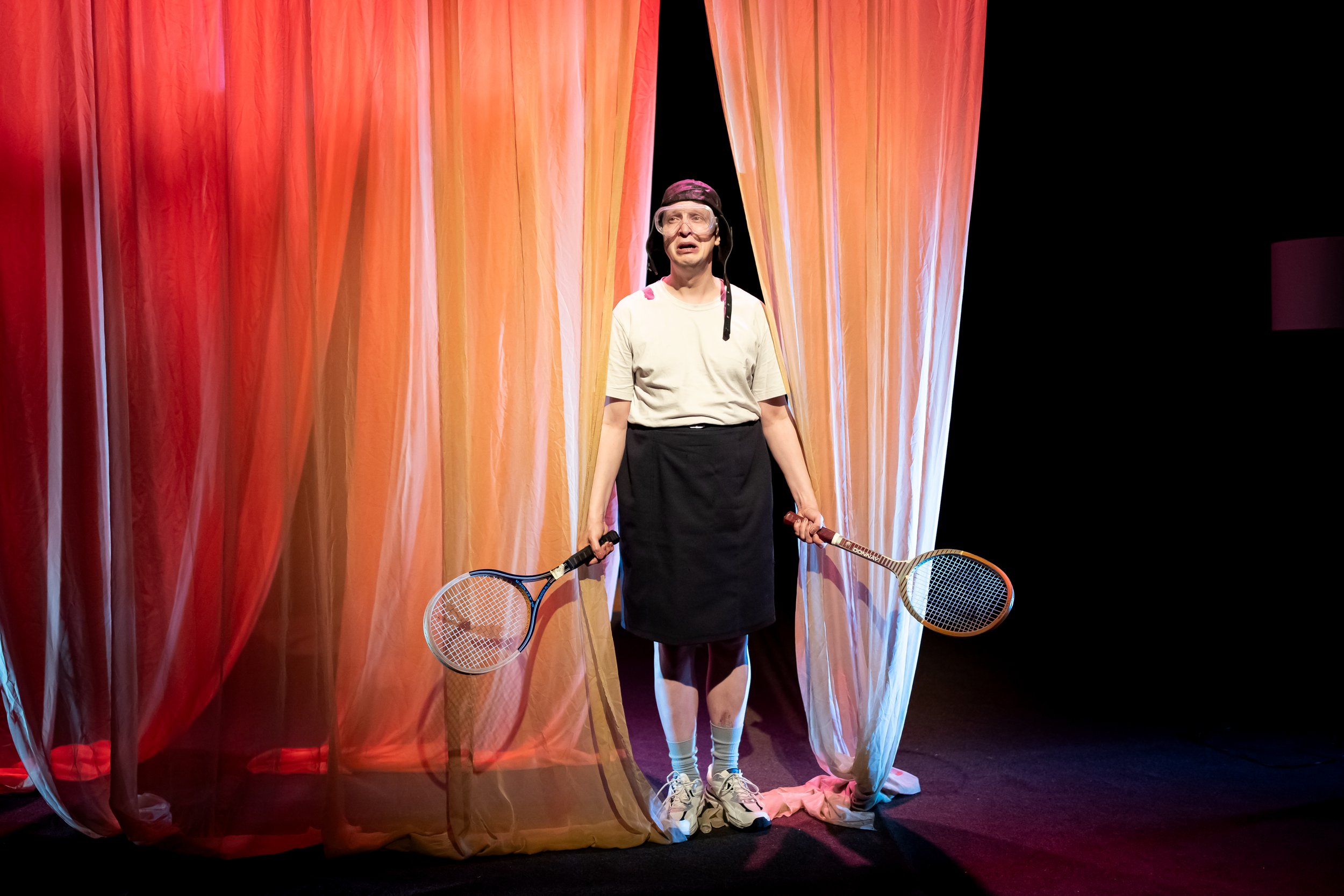 A person stands at a theater curtain holding tennis rackets, dressed in a casual outfit with a white T-shirt, black skirt, and sneakers, with a humorous or theatrical expression.