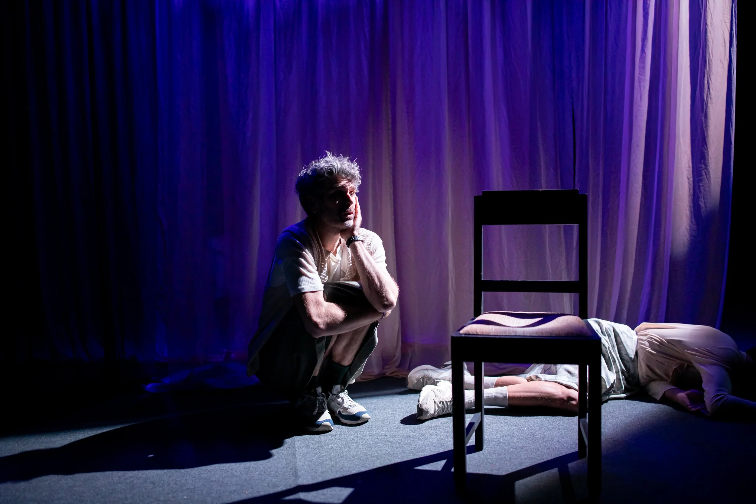 A man crouches next to a person lying face down on the floor, with a chair and a curtain in the background, under dramatic stage lighting.