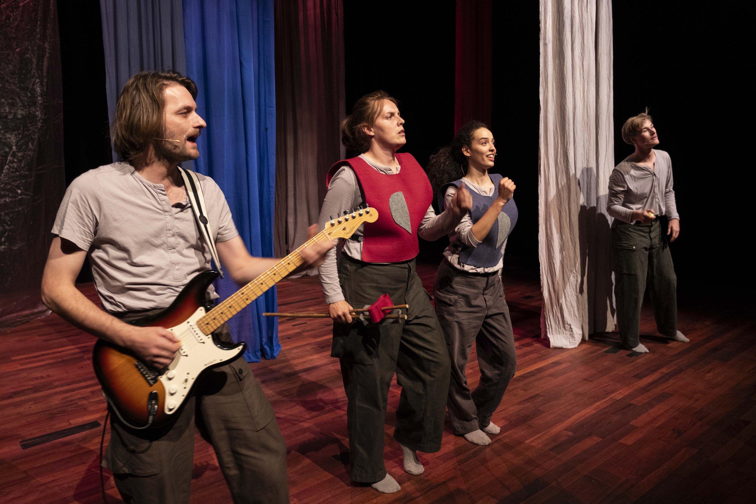 Four actors on stage during a theatrical performance, with colorful curtains in the background. One playing an electric guitar, and the others in expressive poses.