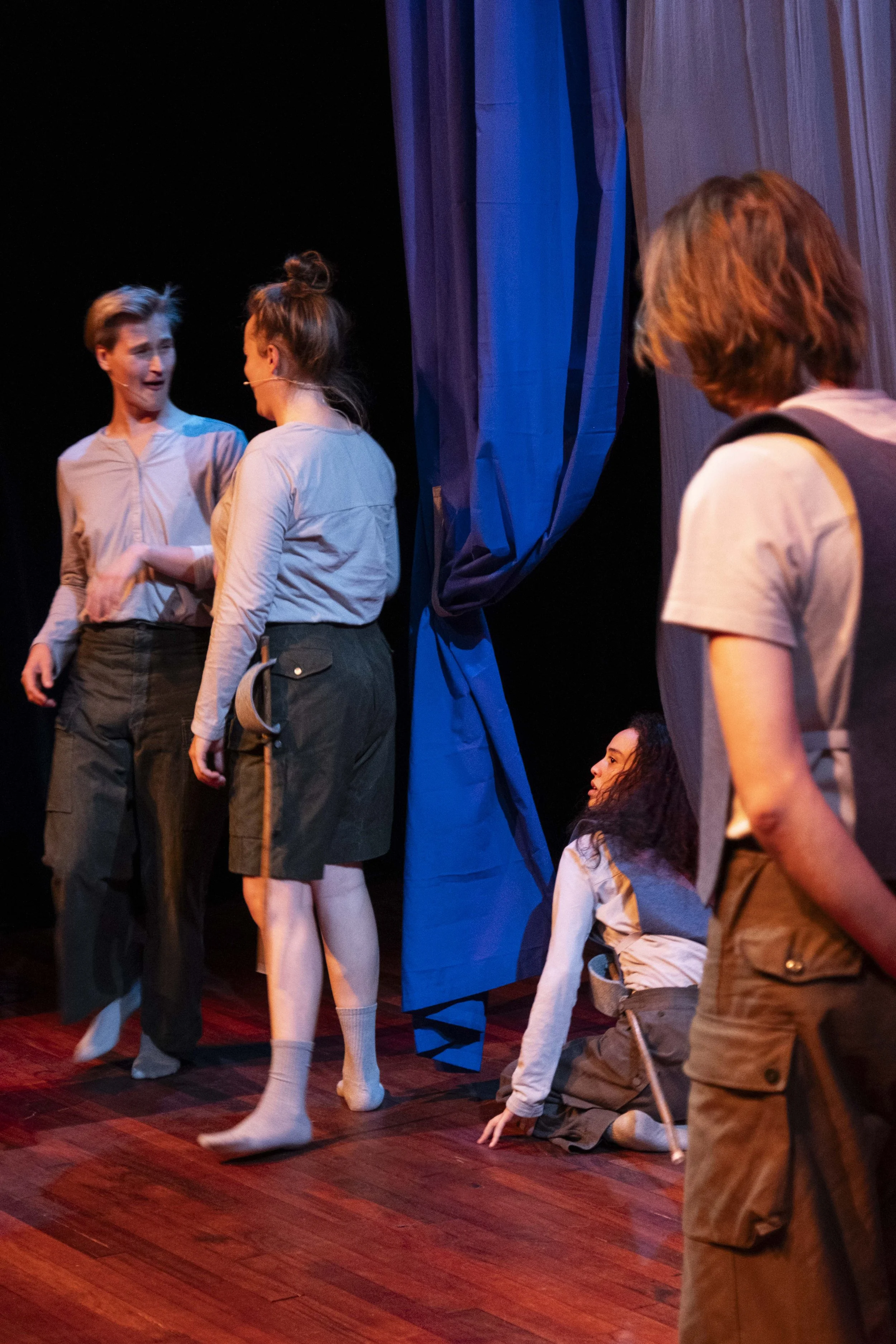 Scene from a stage play with four young actors, two standing and two seated, engaged in a dramatic moment. The stage has wooden floors and blue curtains, with dramatic lighting.