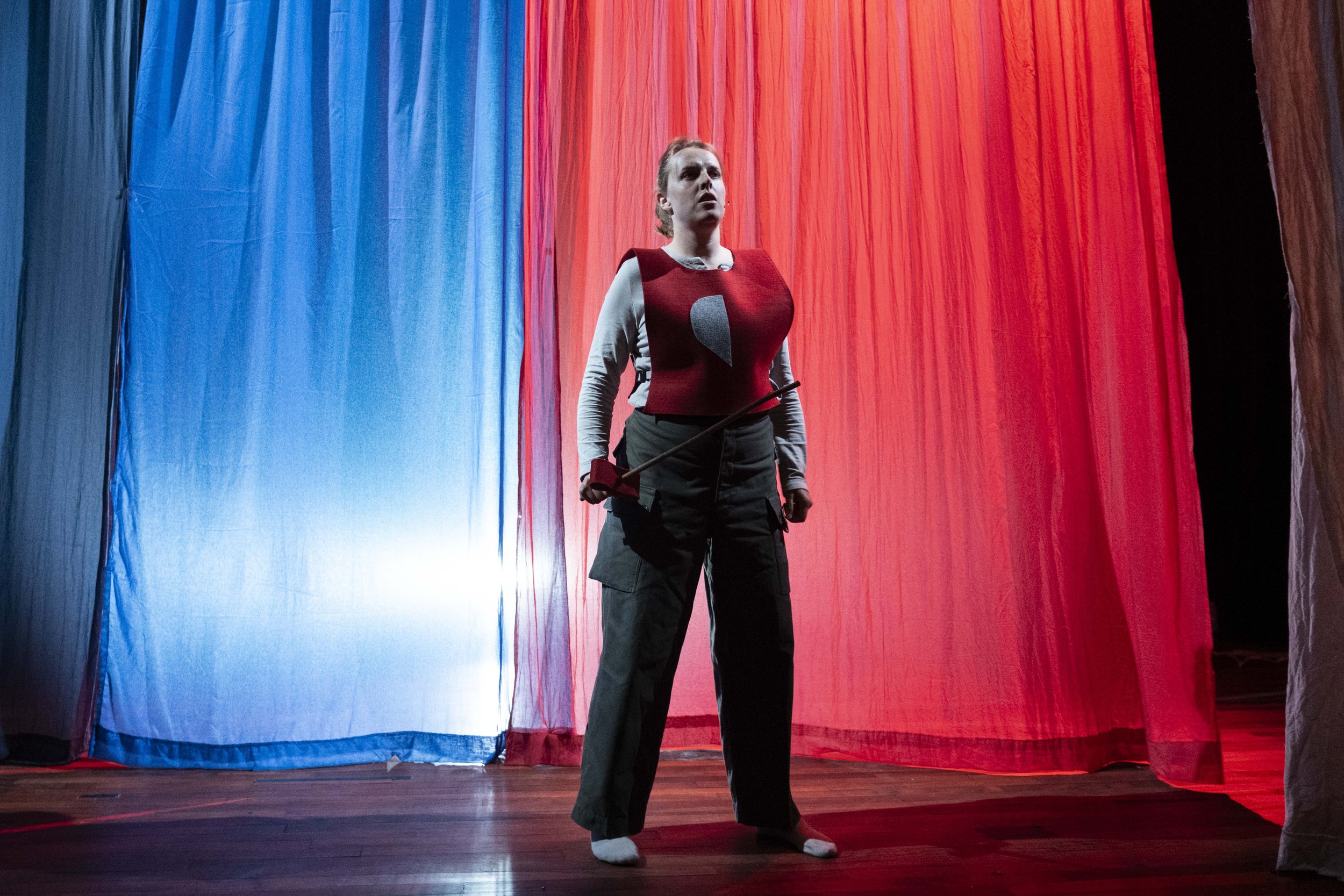 A woman stands on a stage with red, blue, and black curtains, dressed in casual pants, long-sleeve shirt, and a red vest, holding a fencing foil.