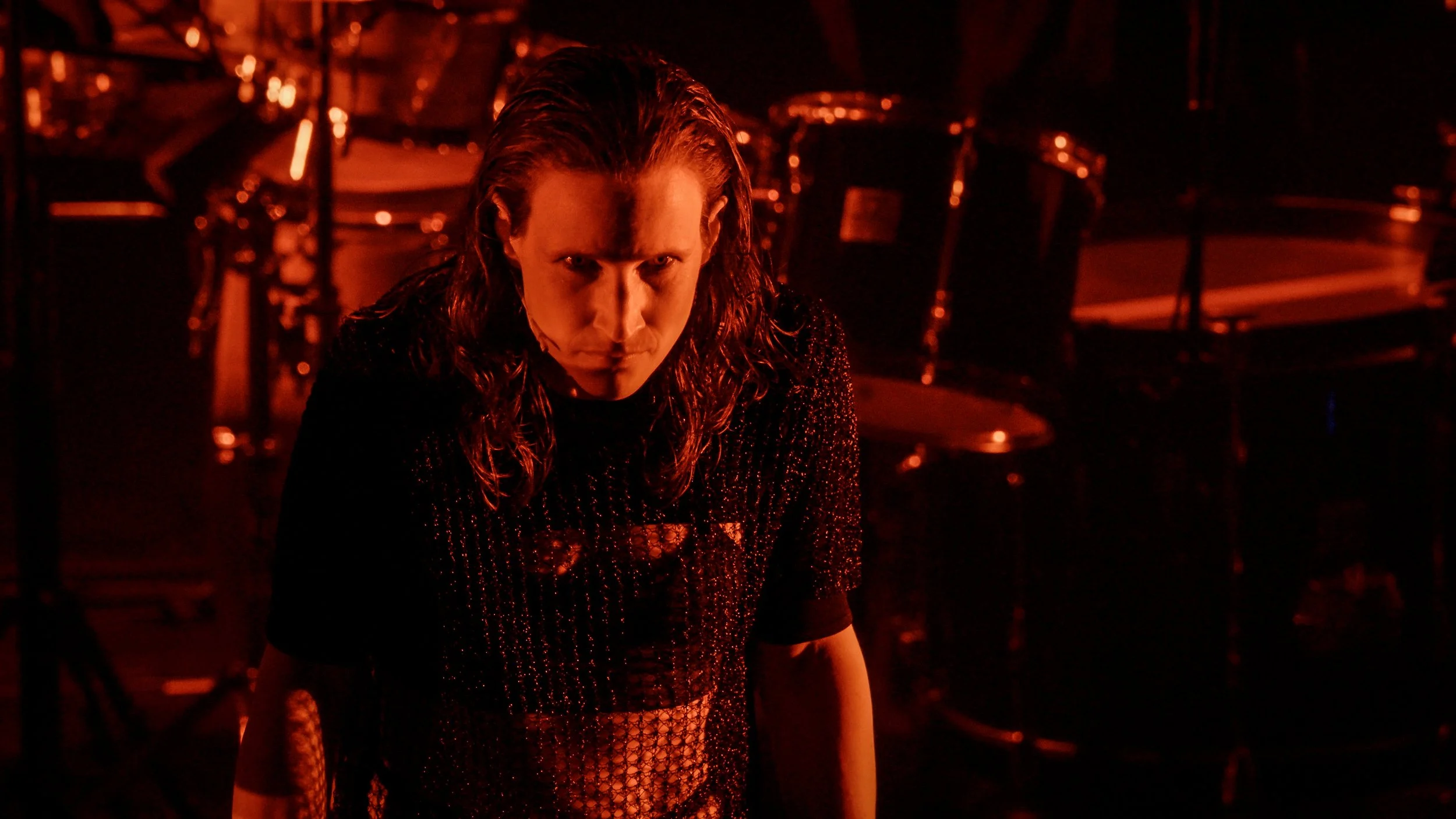 A person with long, wet hair looking intensely at the camera, surrounded by dim red lighting and empty chairs, with drums in the background.
