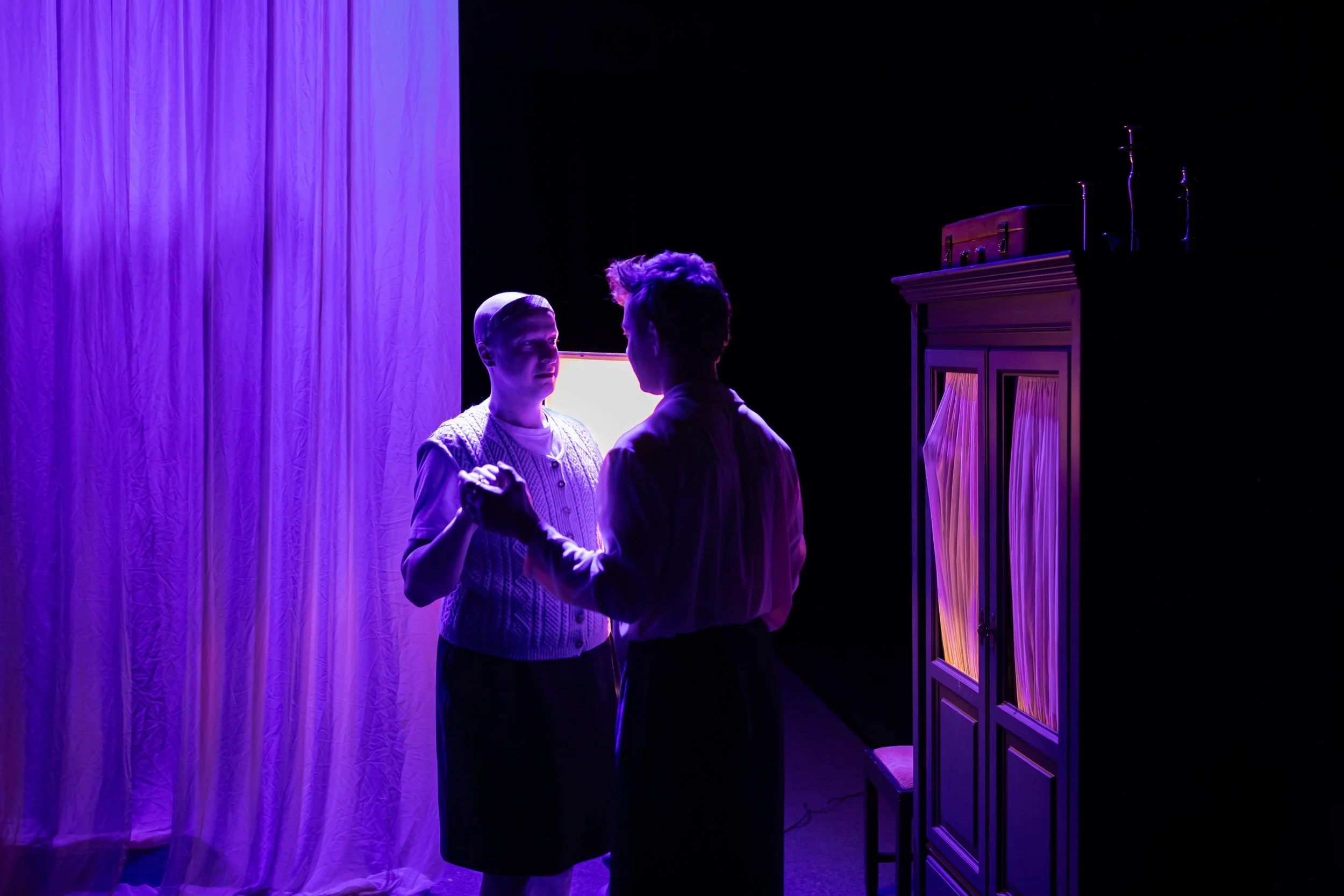 Two actors on stage under purple lighting, holding hands and gazing at each other, with a purple curtain on the left and a cabinet with curtains on the right.