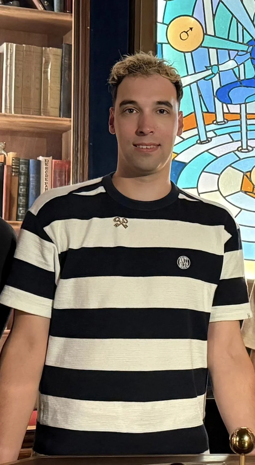 A young man with short, curly hair, wearing a black and white horizontal striped t-shirt with small metal pins on the collar, standing indoors near a colorful stained-glass window with mathematical symbols and science equipment designs, and a wooden bookshelf filled with books behind him.