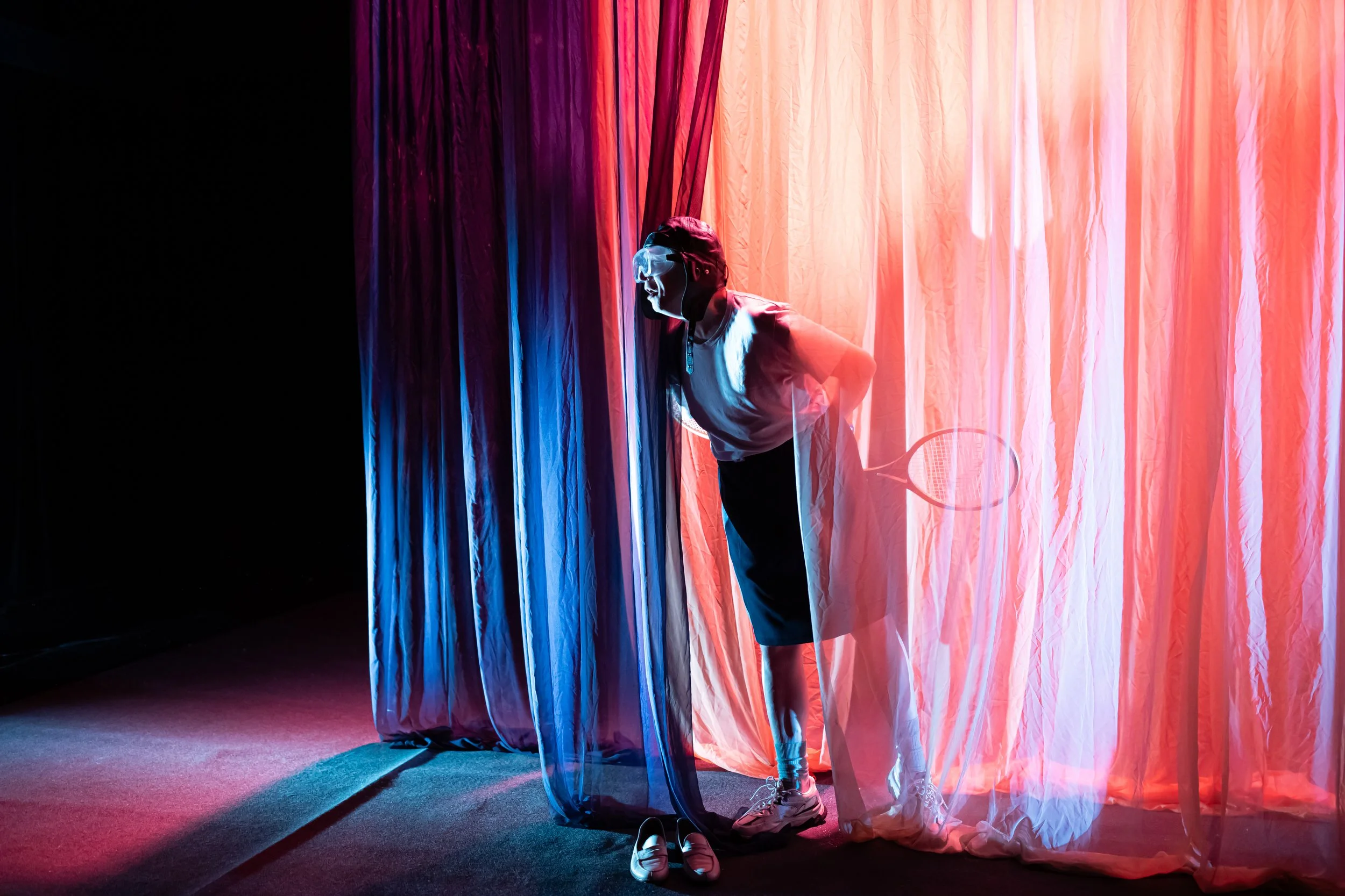 Person holding a tennis racket peering out from behind pink and purple curtains in a dimly lit room.