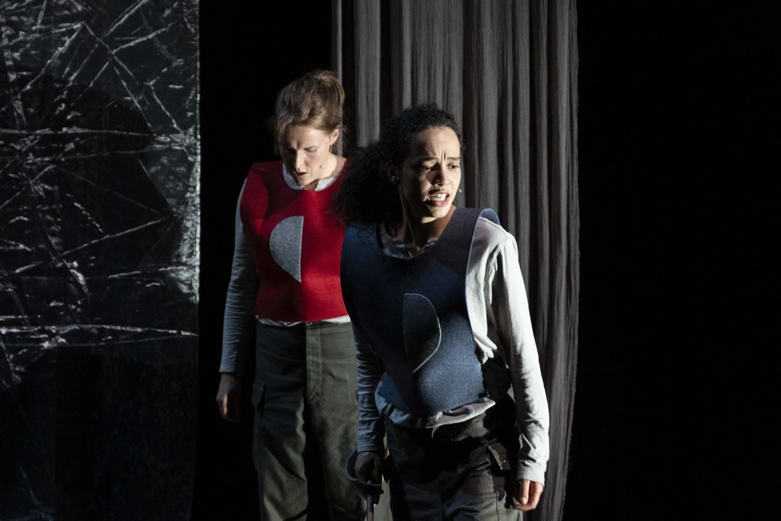 Two women on stage with distressed expressions, wearing colorful mismatched costumes and standing in front of dark, textured backgrounds.