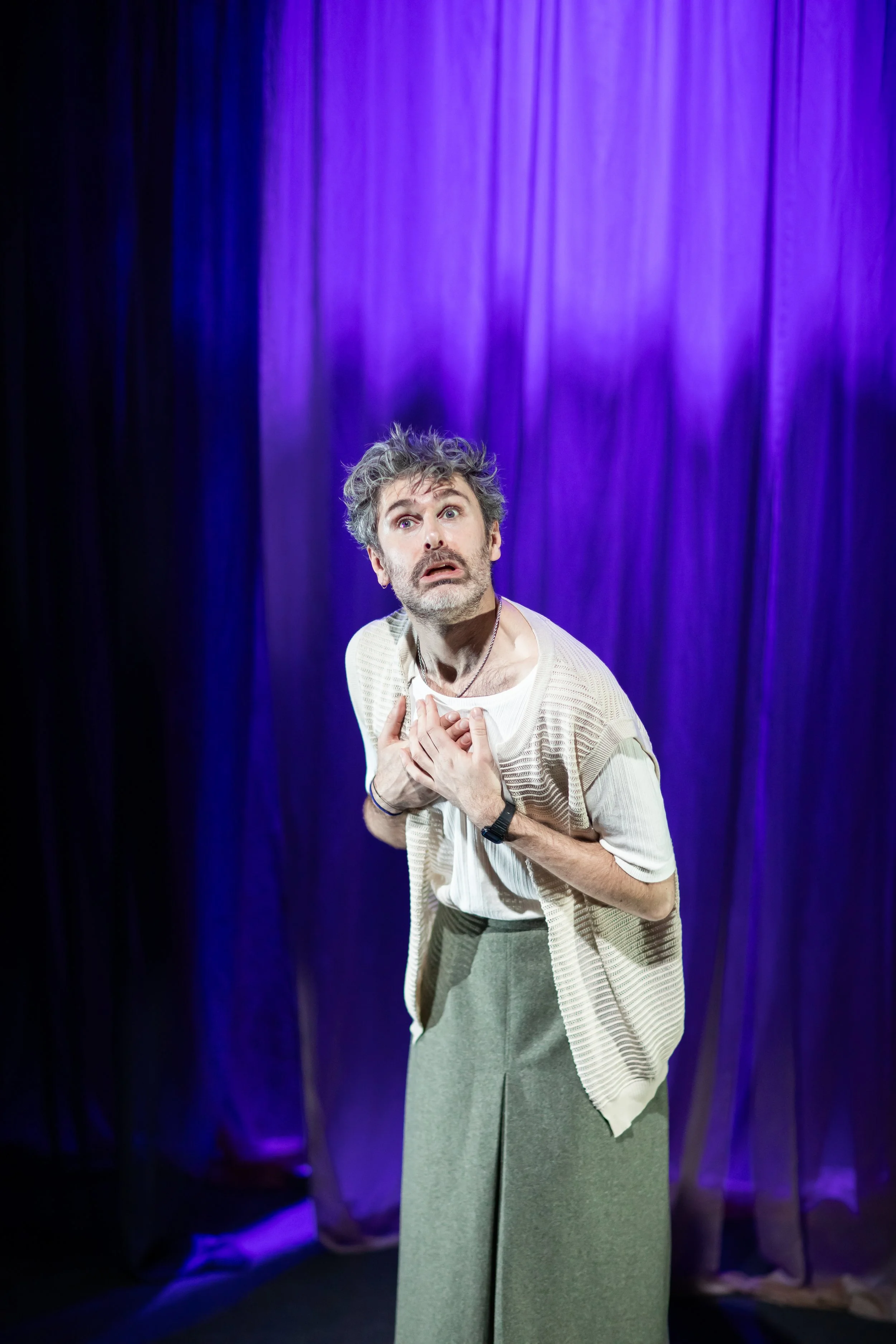 Man with gray hair and beard, wearing a beige sweater vest and loose pants, performing on stage with purple curtains in the background, holding his chest and looking surprised or emotional.