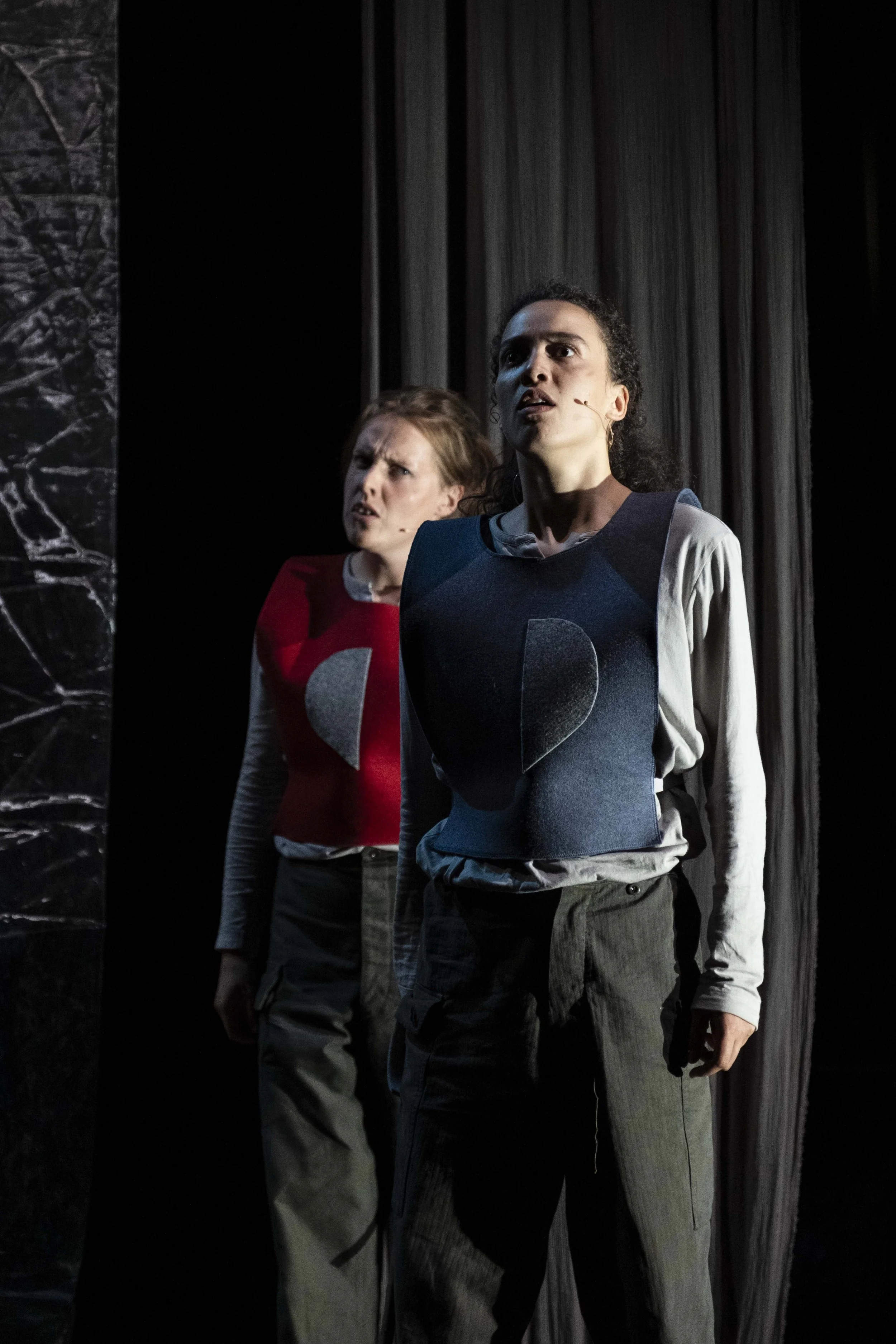 Two women on stage, one in a red vest and the other in a black vest, both showing serious or emotional expressions.