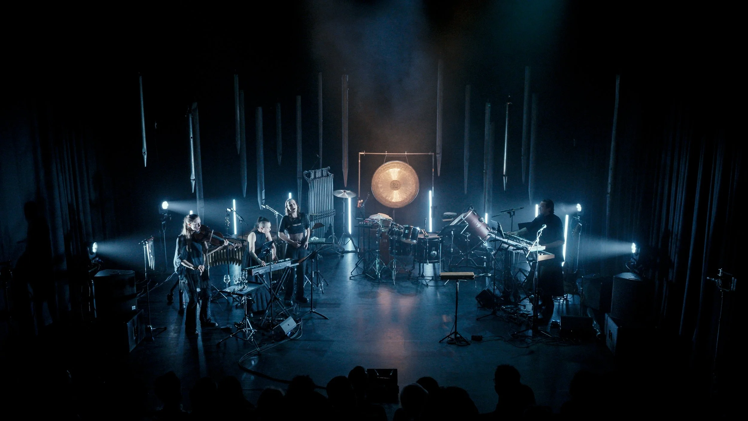 A musical performance on stage featuring a drummer, a keyboardist, two violinists, and a vibraphone player, with stage lighting and a gong in the background.
