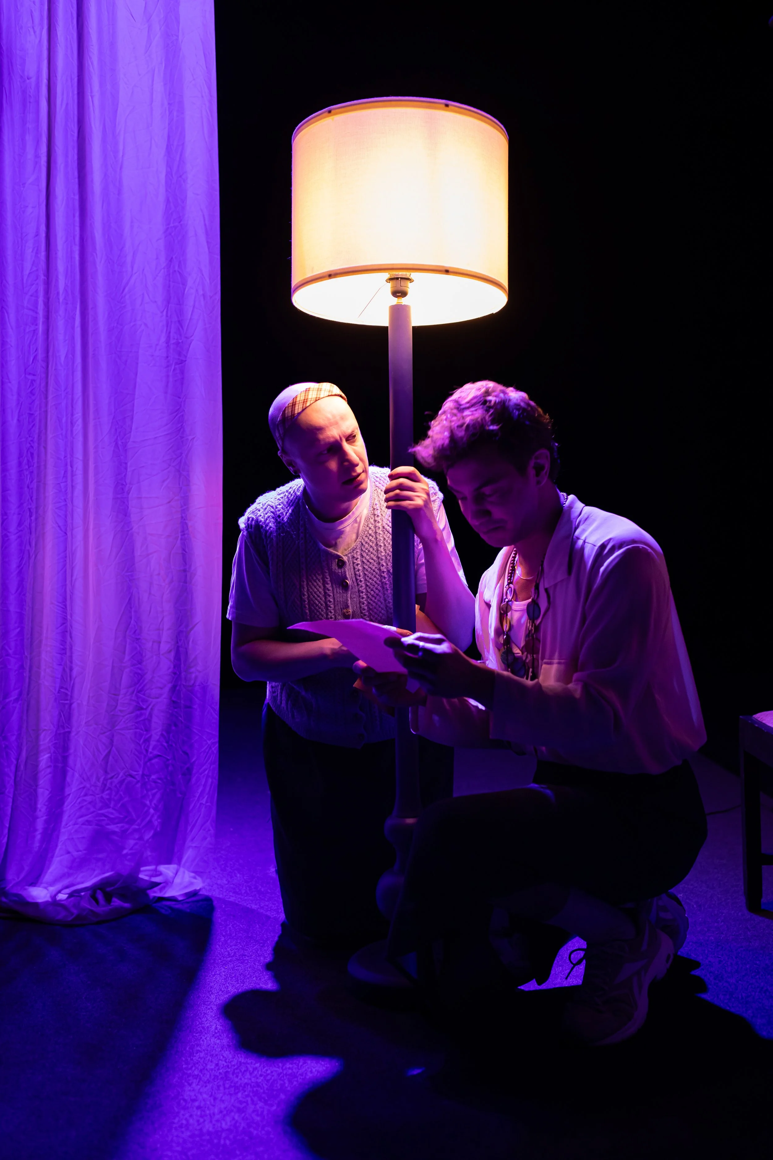 Two actors performing on stage under a large standing lampshade, with purple lighting. One actor is sitting, reading from a script, and the other is standing beside, looking at the script.