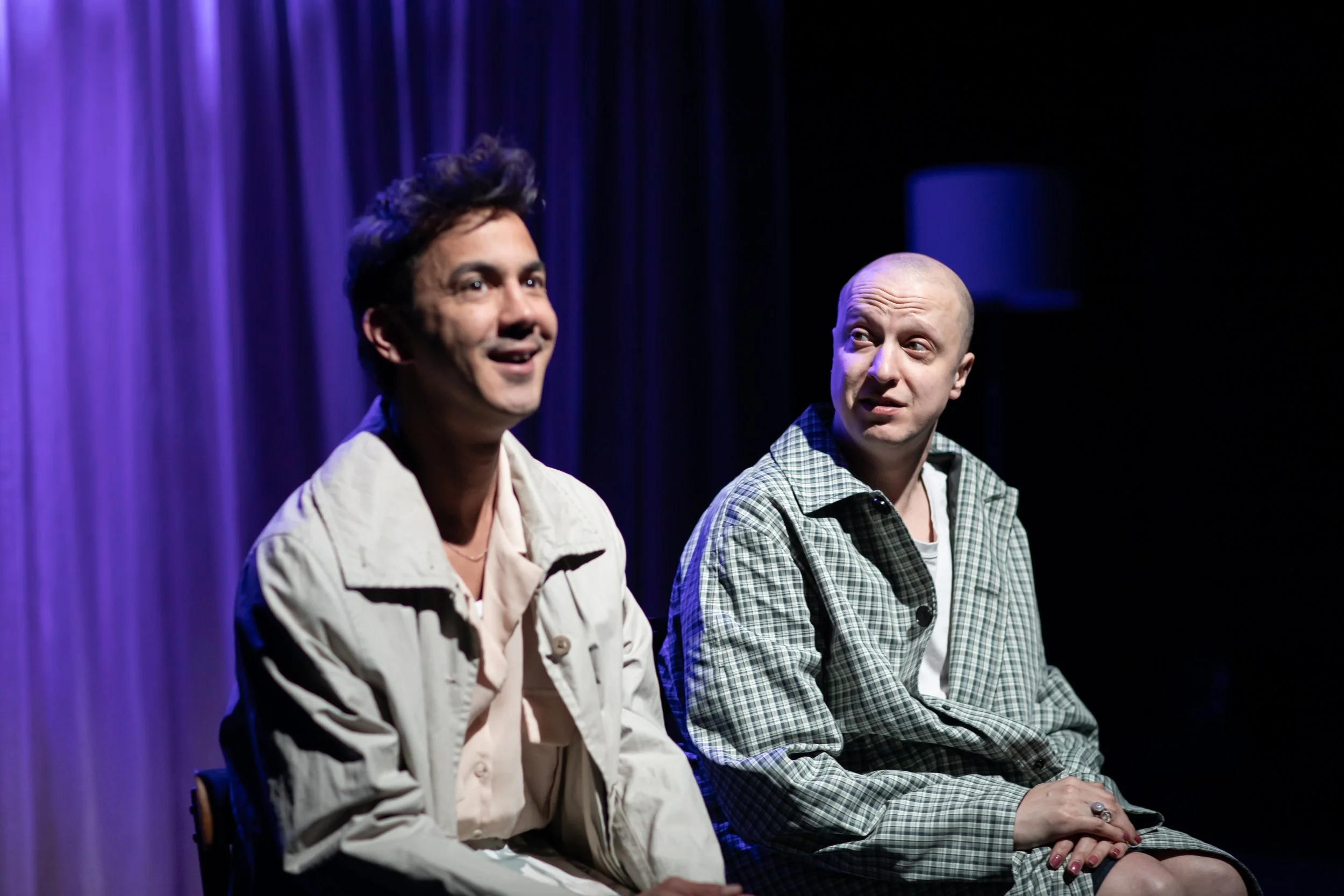 Two men sitting on a stage with purple curtains, engaged in conversation. The man on the left has dark hair and is smiling, wearing a light-colored jacket. The man on the right is bald, listening attentively, wearing a checkered jacket.