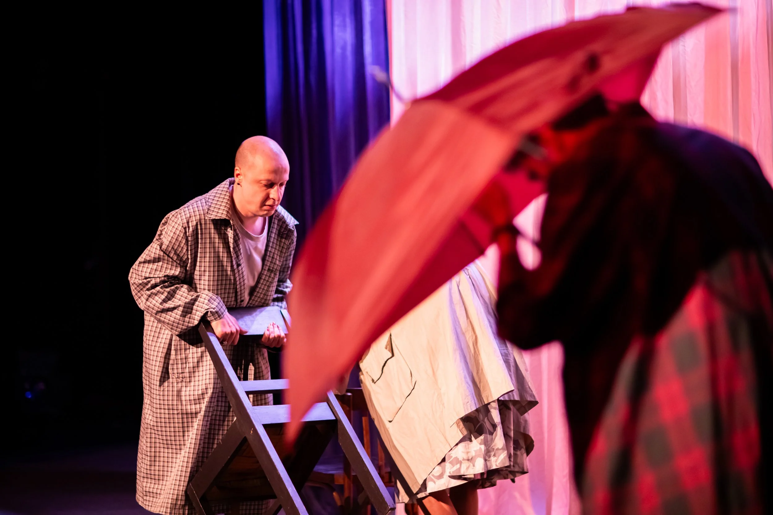 A man in a checkered coat stands on stage behind a wooden chair, looking down. In the foreground, a person wearing a red hood and plaid shirt is partially visible, facing the stage. The background features stage curtains with pink and purple lighting