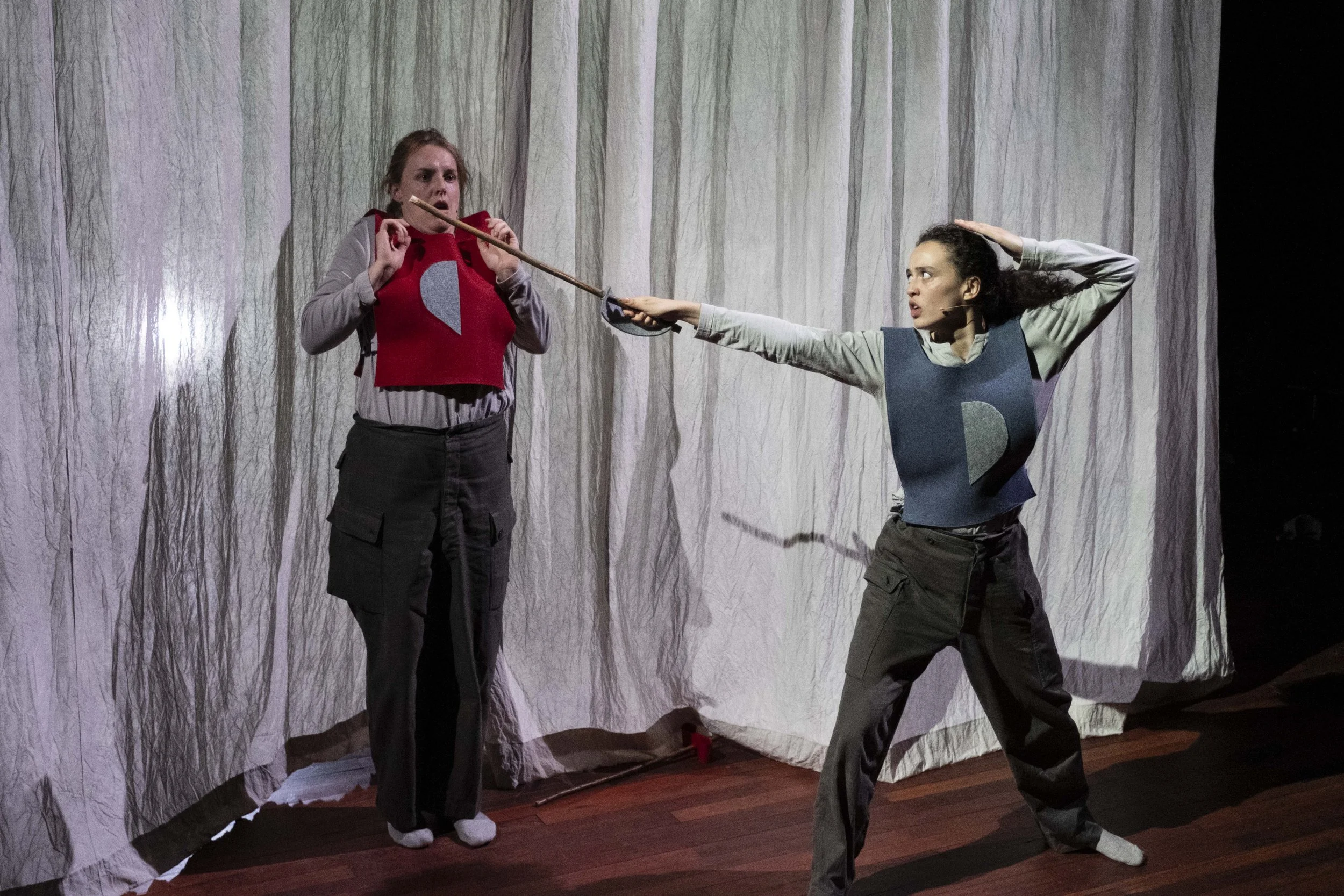 Two women in costume on stage, one with a spear-like object pointed at the other, with a backdrop resembling a forest scene.