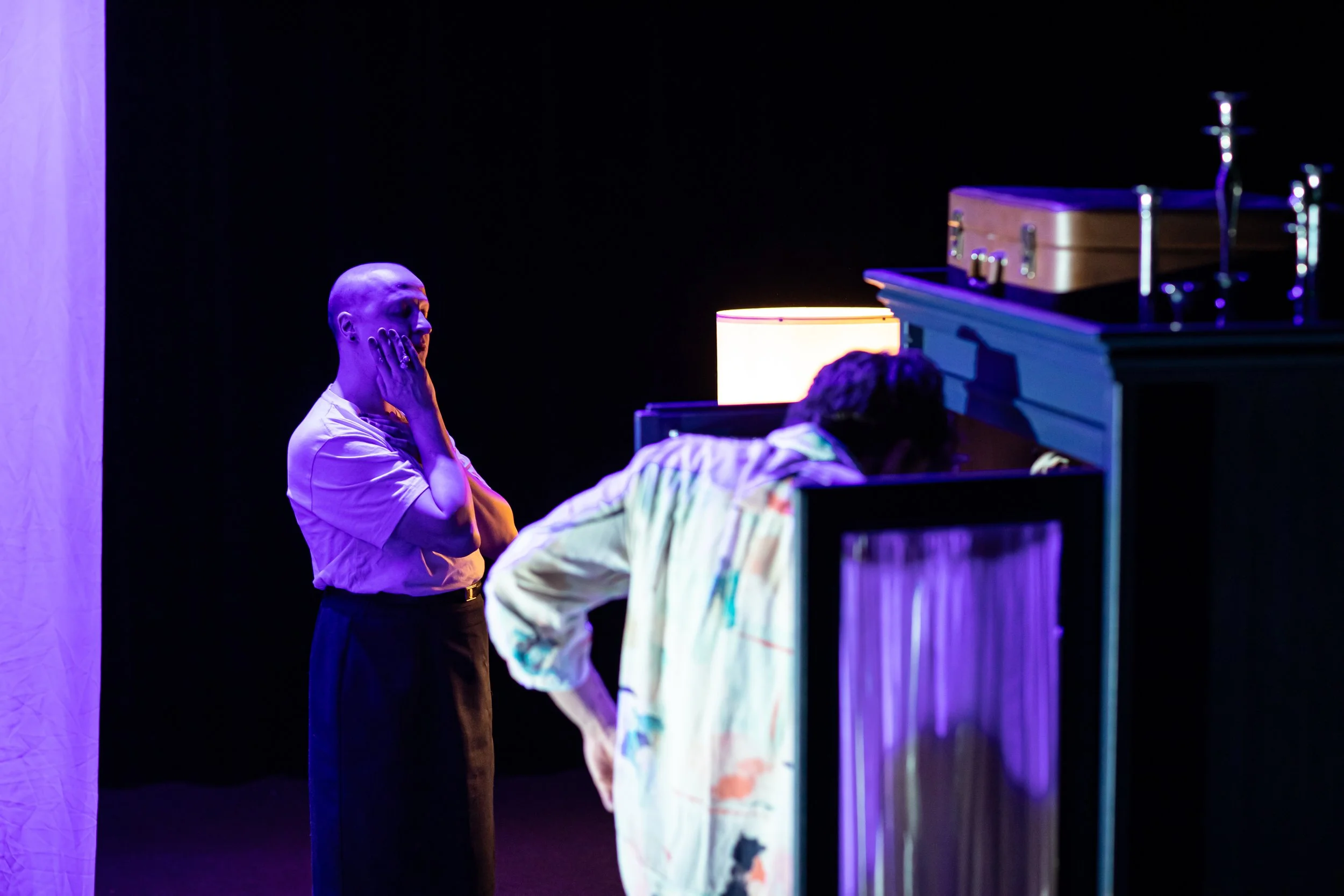 Two individuals on stage under purple lighting, one with hands on face and eyes closed, the other bent over a table or desk, with a lamp and a dragon figurine in the background.