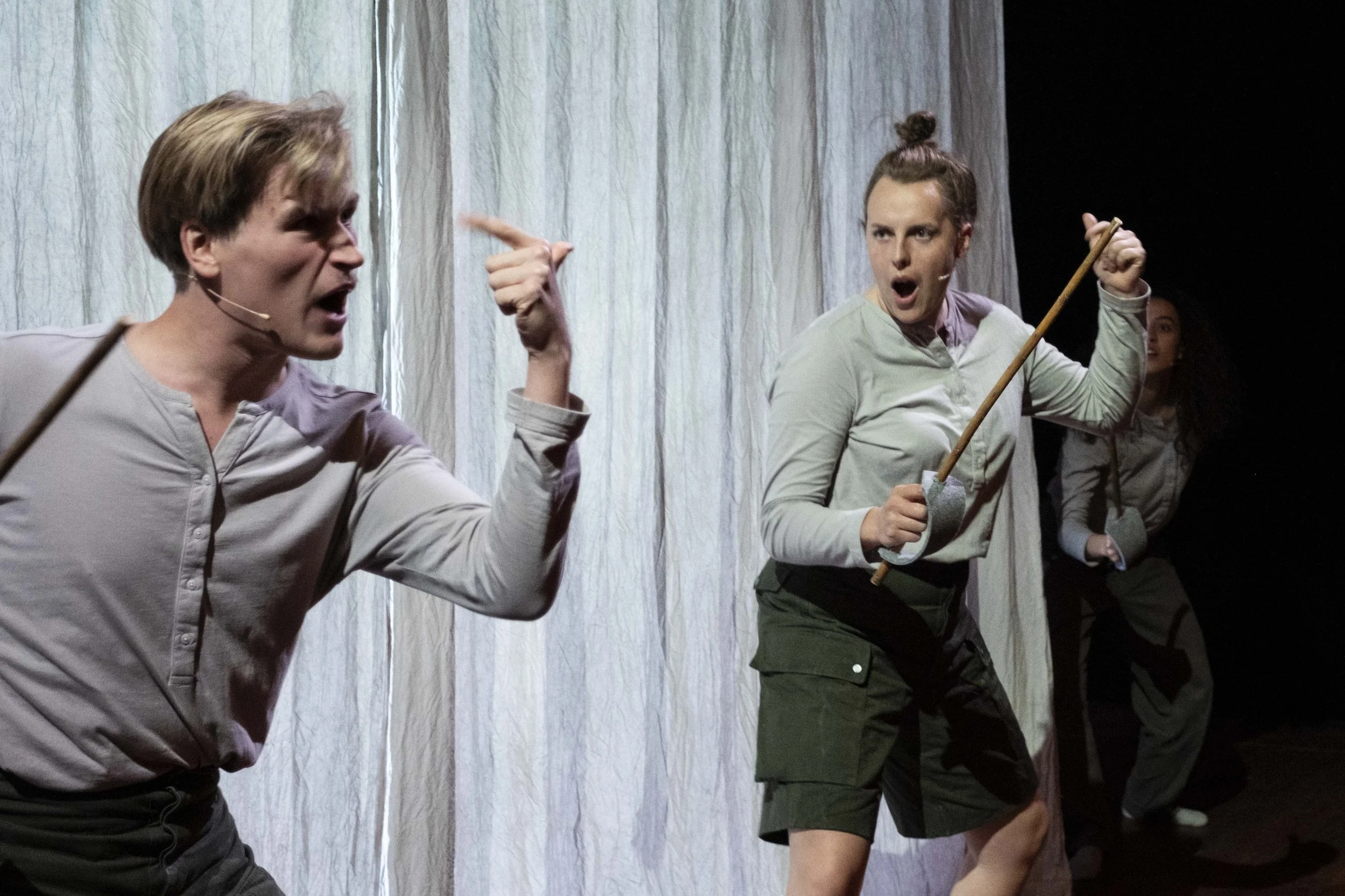 Three actors on stage, engaged in a theatrical performance. The actor on the left is gesturing passionately, the middle actor is holding a stick and appears to be yelling, and the actress in the back is partially visible. The stage has a white curtai