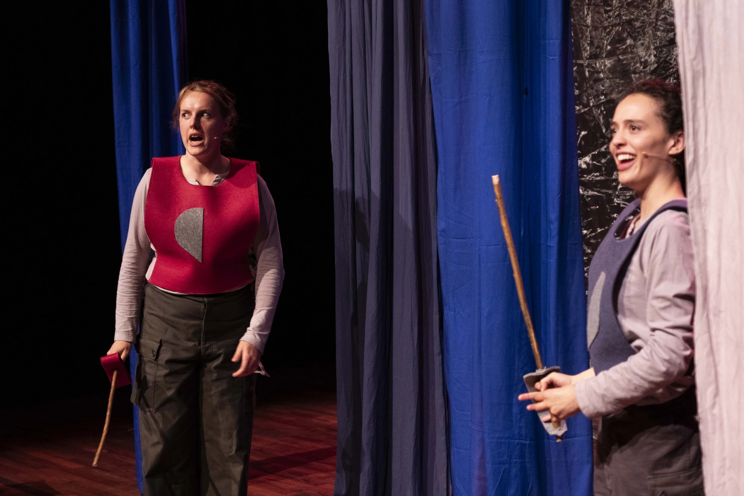 Two women on stage, engaged in a theatrical performance. One woman stands on the left, wearing a red vest with a gray design, gray long-sleeve shirt, and dark cargo pants, holding a stick. The other woman stands on the right, partially behind a curta