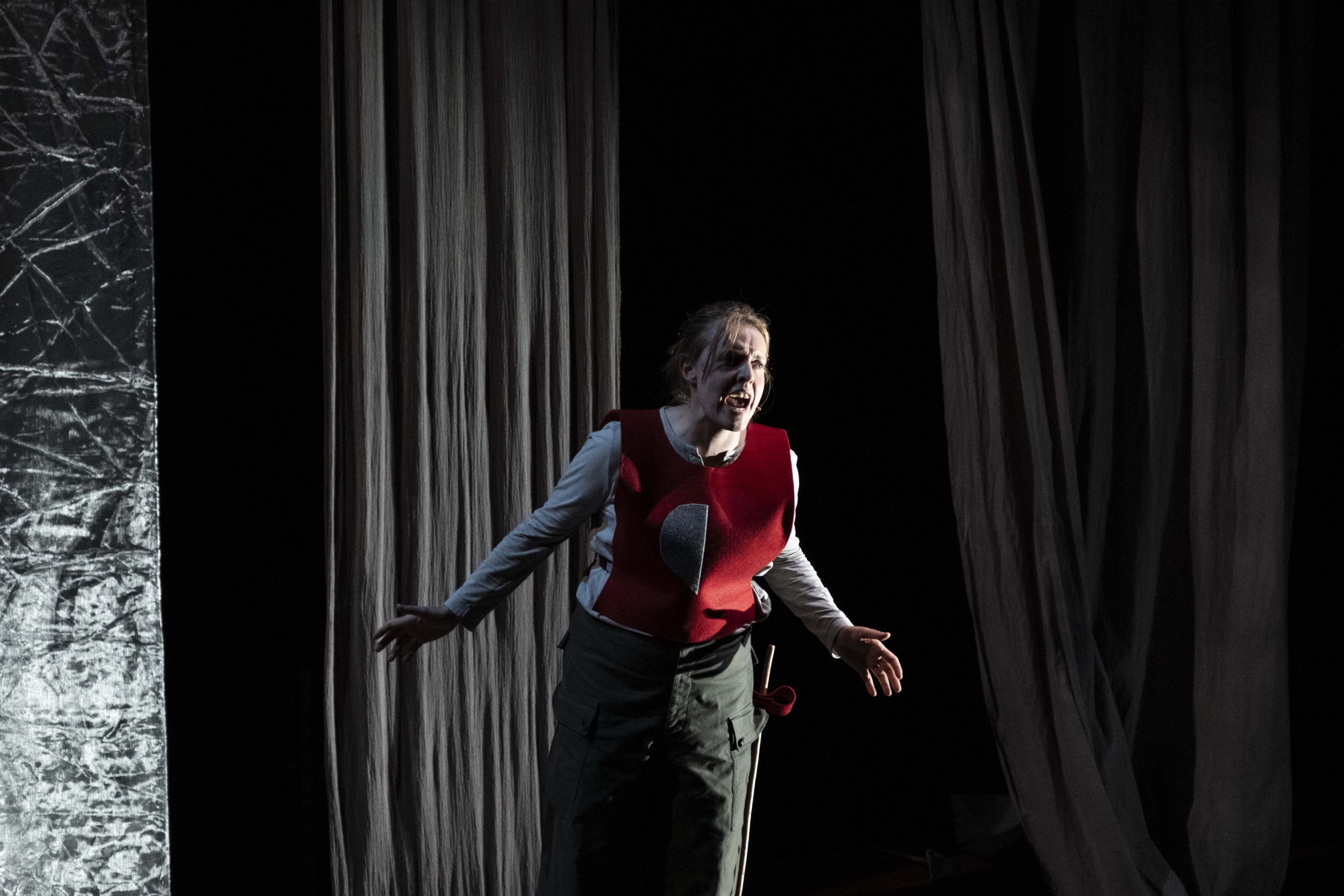 A woman performing on stage, expressing emotion with her mouth open and arms slightly outstretched, with curtains and stage lighting in the background.
