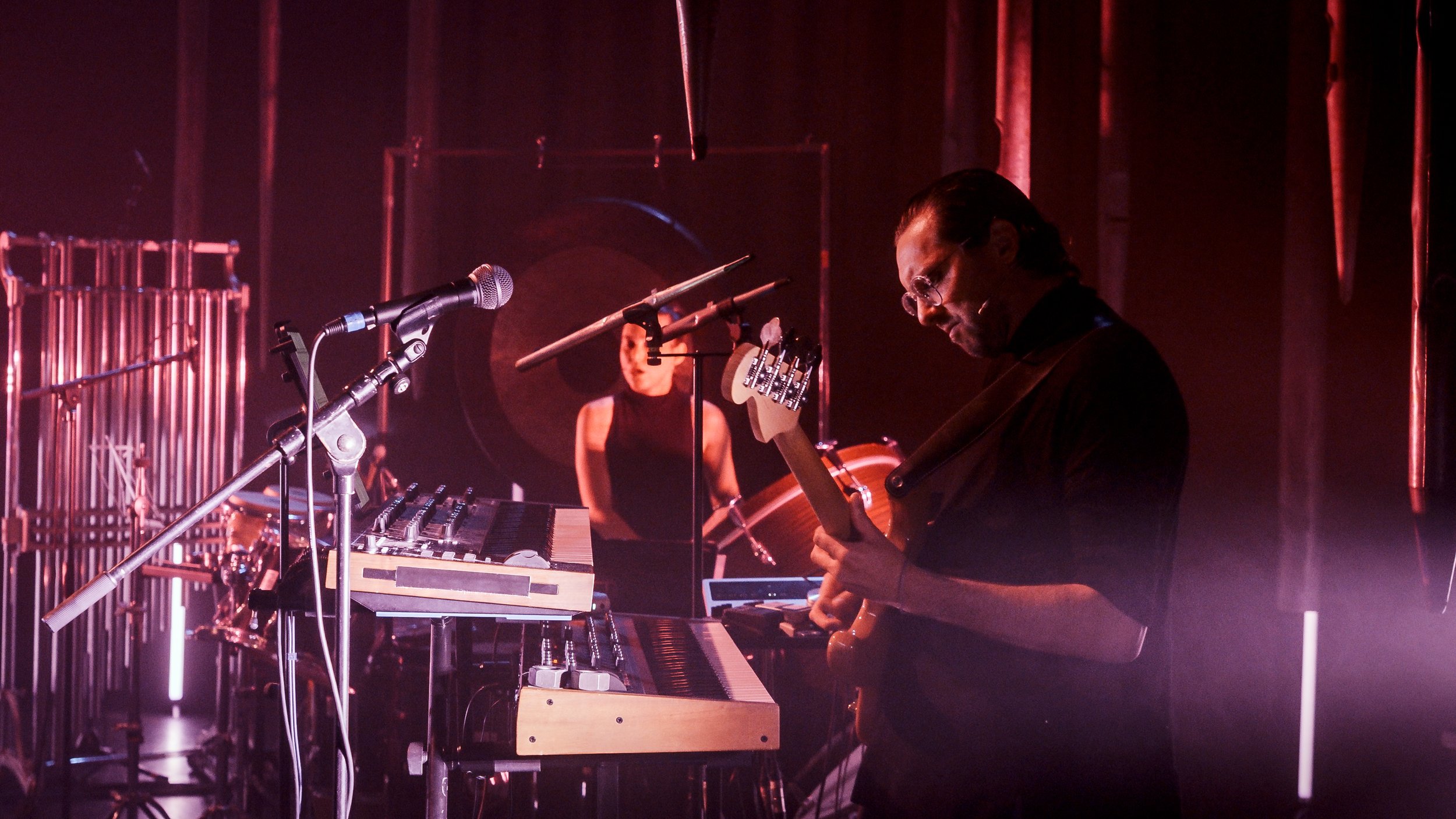 A musician playing the guitar and a drummer performing on stage with electronic music equipment under purple and pink stage lighting.