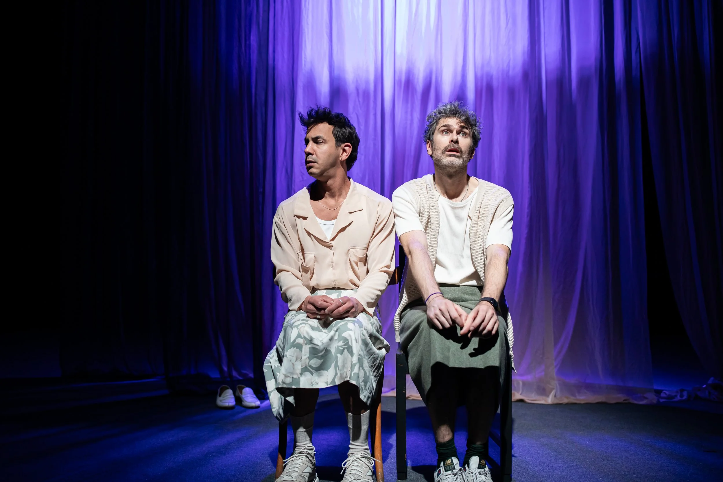 Two men sitting on chairs on stage with purple curtain background, both looking upward with thoughtful expressions, dressed casually in light-colored clothing.