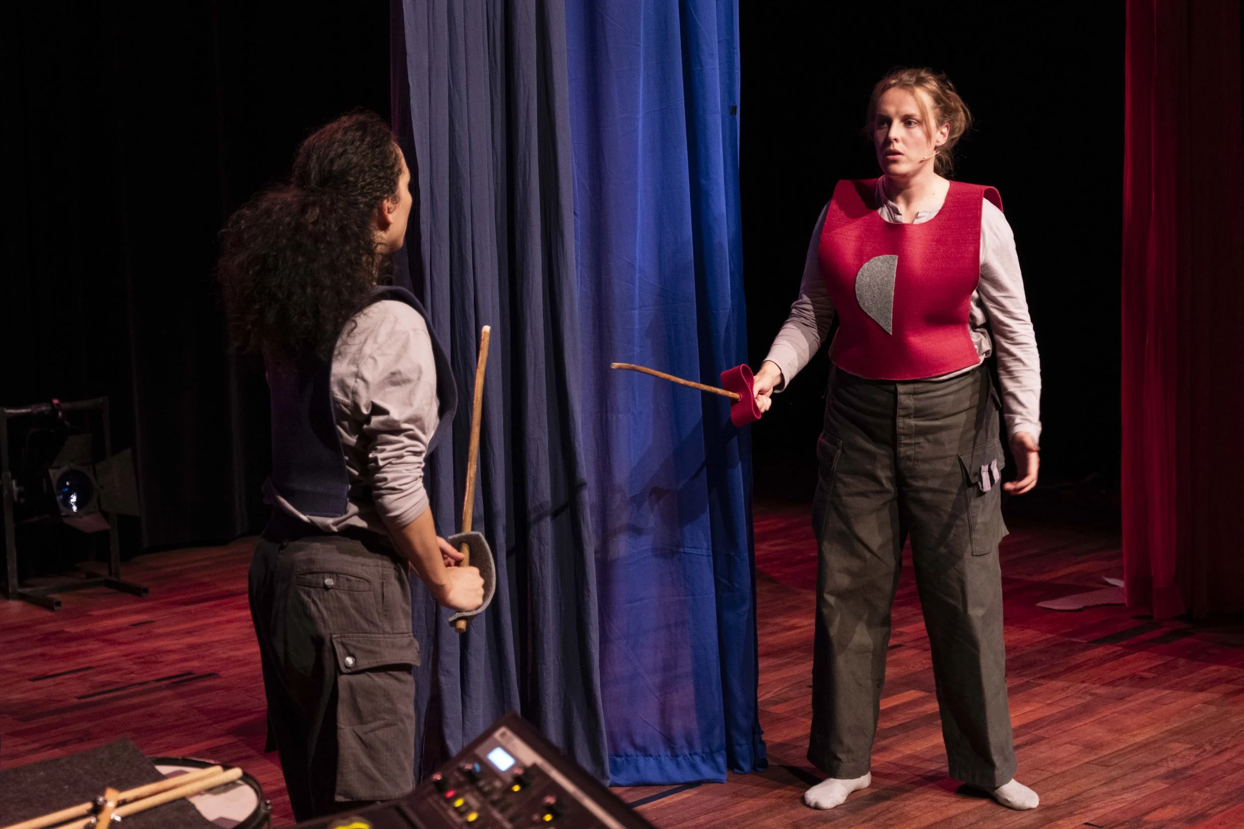 Two women on stage holding sticks and engaging in a dramatic scene, with a curtain in the background.