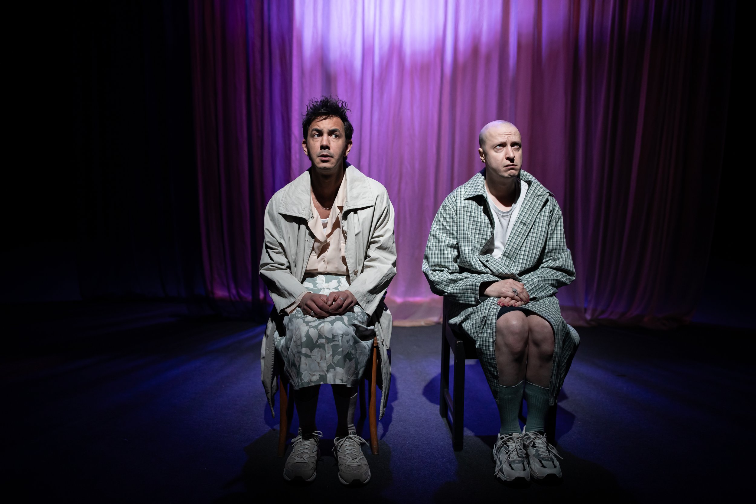Two men sitting on chairs on a stage with purple curtains behind them, both wearing casual clothing and looking in different directions.
