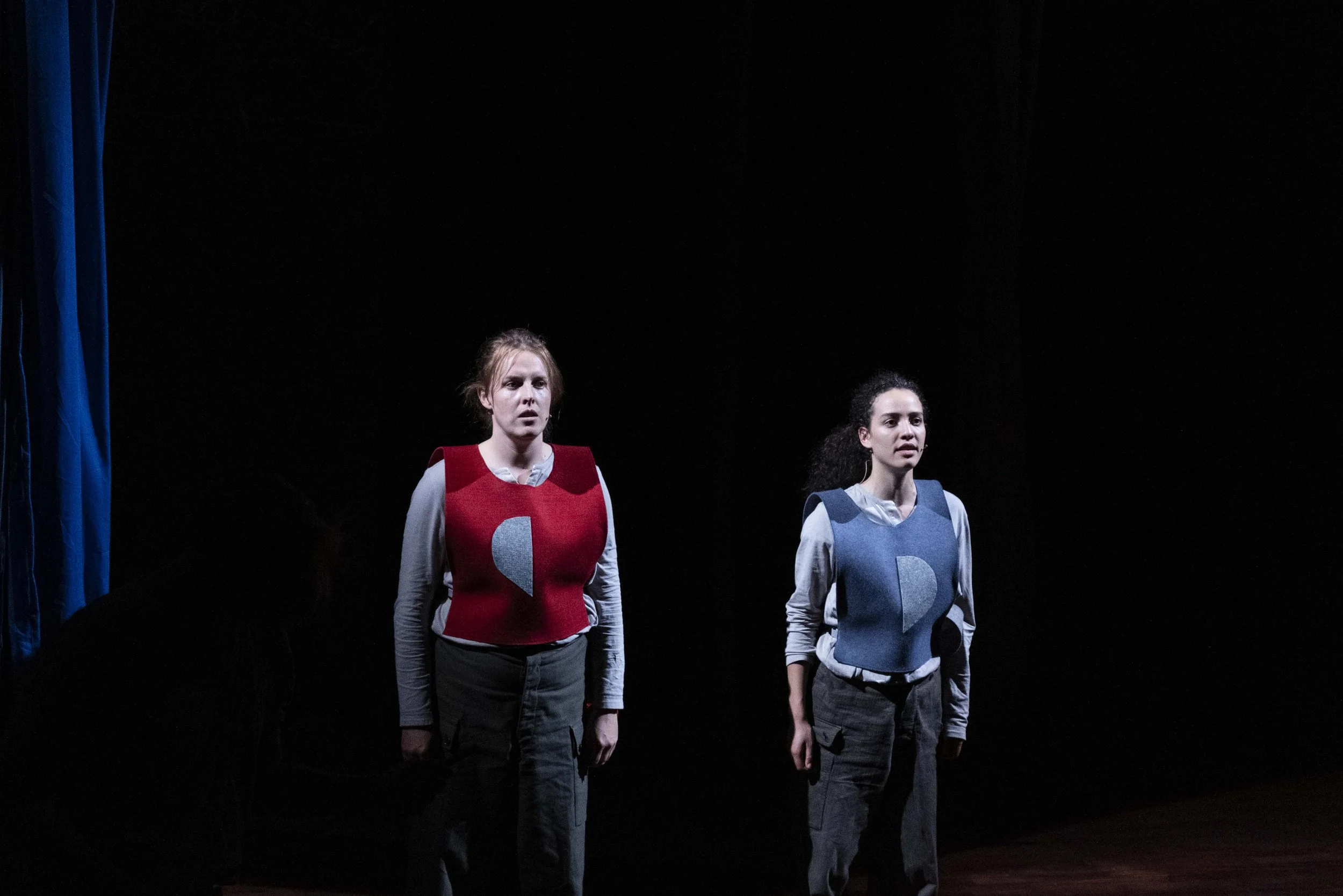 Two women on stage, wearing vests with half moon designs, standing in a dark theater with a blue curtain on the left.