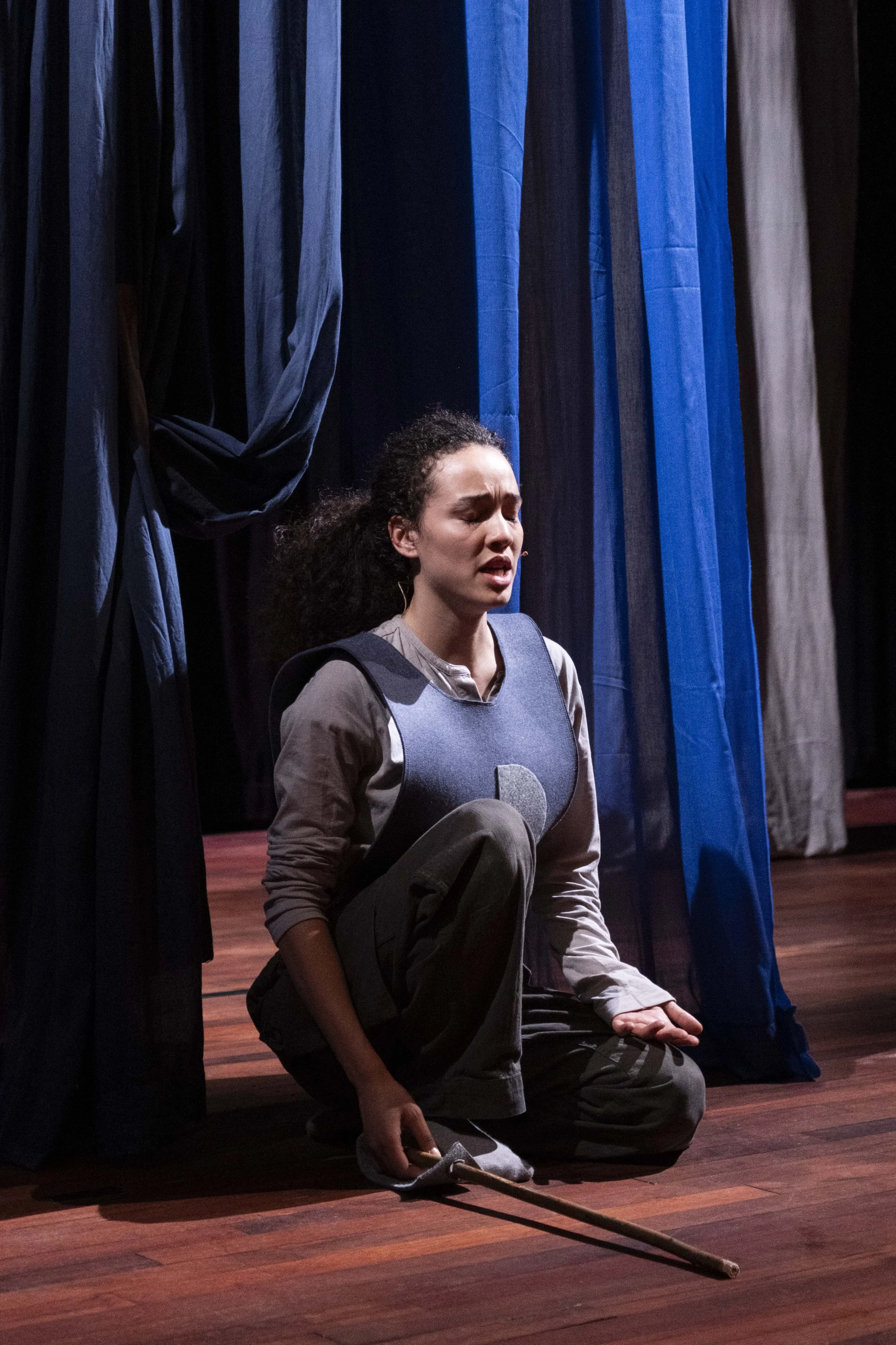 A woman on stage, kneeling with a focused and emotional expression, surrounded by blue stage curtains, holding a wooden stick.