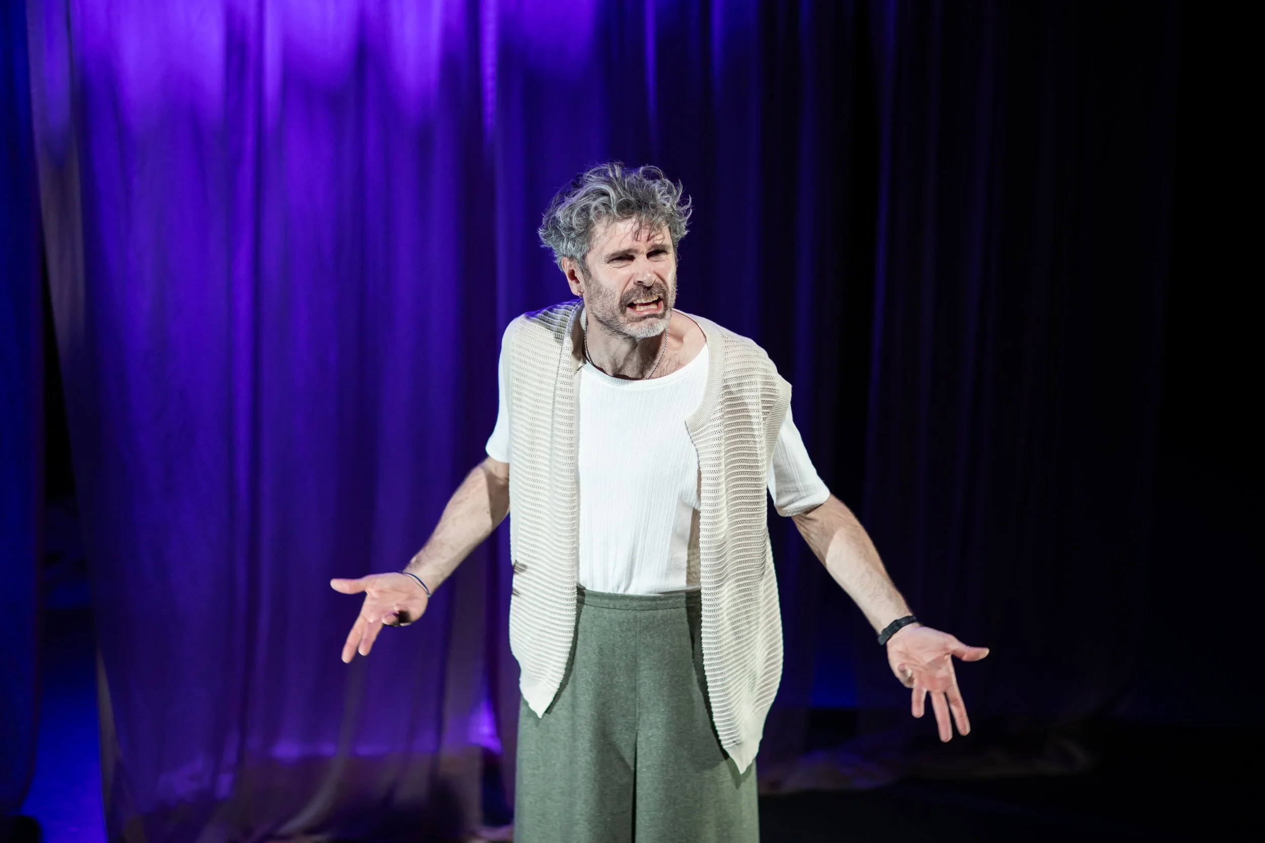 A man with gray curly hair and beard, wearing a white shirt and beige vest, standing on a stage with purple curtains in the background, gesturing with his hands and speaking.