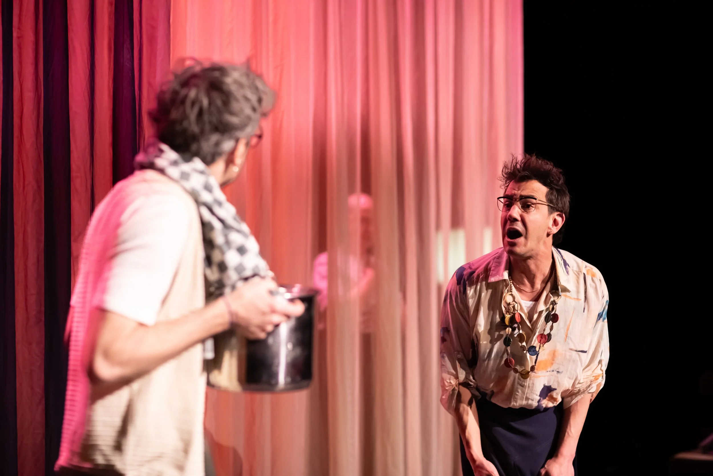 Two women having a conversation on stage, one with glasses and a necklace, the other holding a cup, with pink curtains in the background.