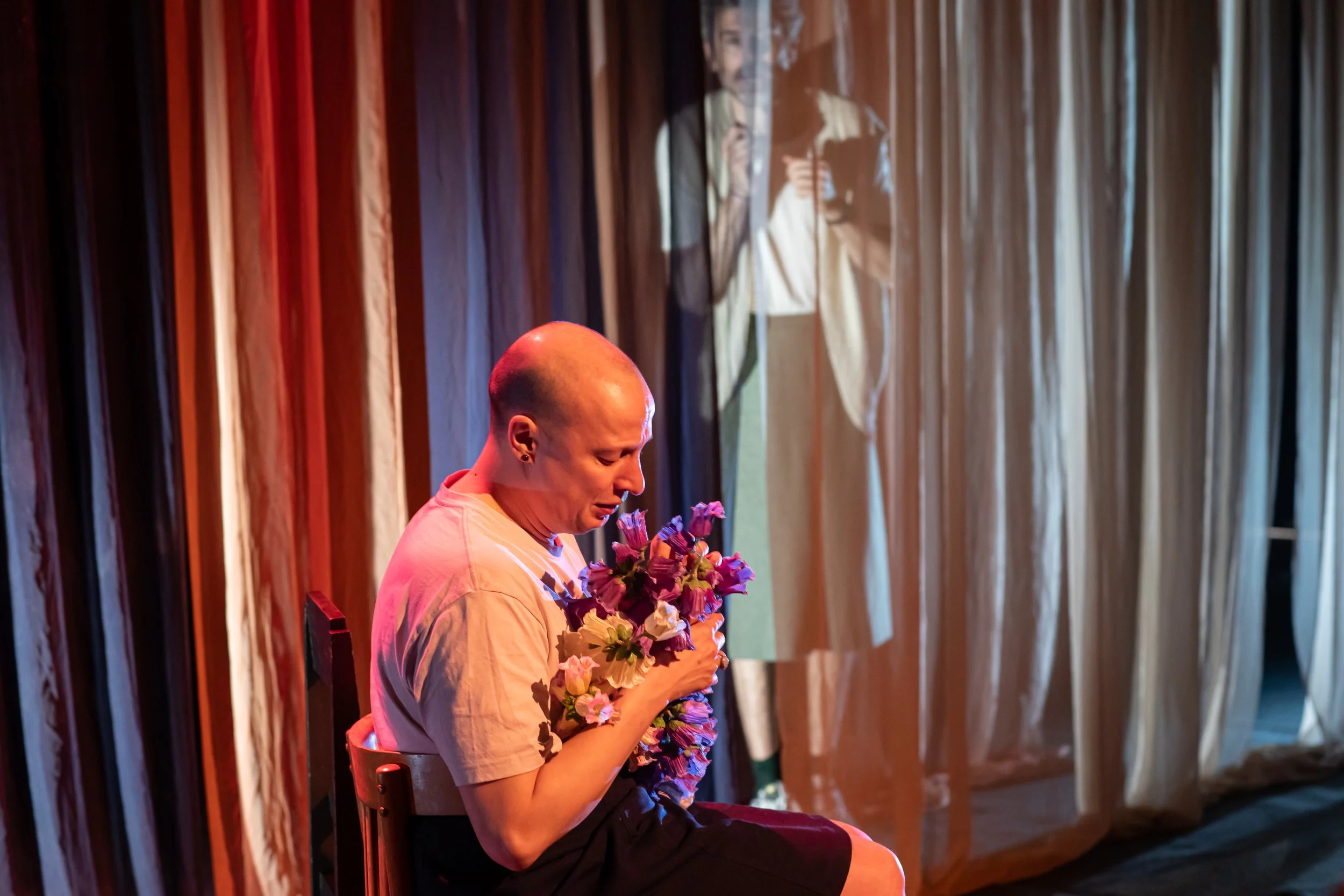 A person with a shaved head sitting on a chair, holding a bouquet of pink and purple flowers, with a backdrop of fabric curtains. An image of another person taking a photo is visible in the semi-transparent curtain.