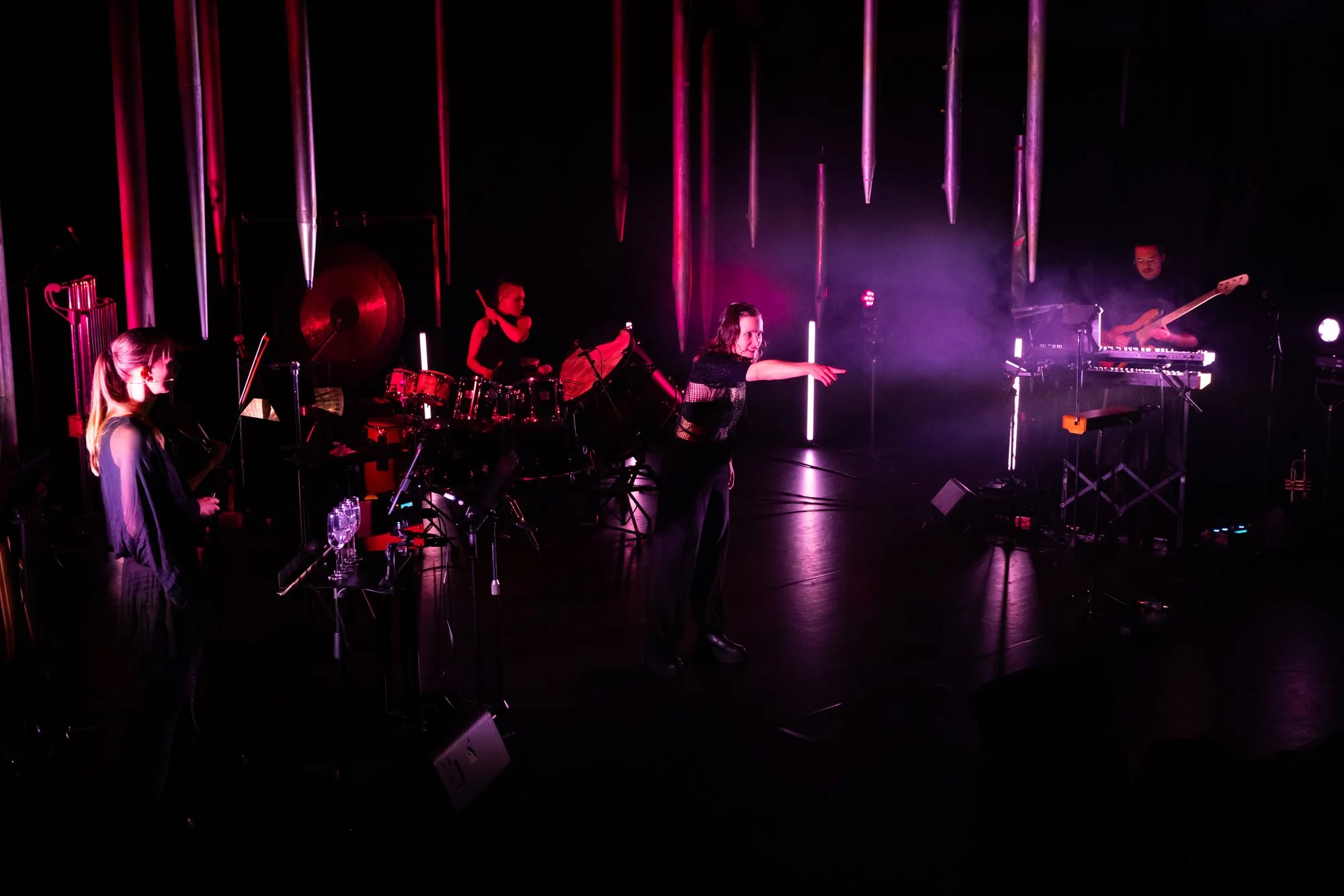 Musicians performing on stage with pink and purple lighting, including a woman conducting, a violinist, a drummer, and a keyboardist, with vertical metallic tubes in the background.