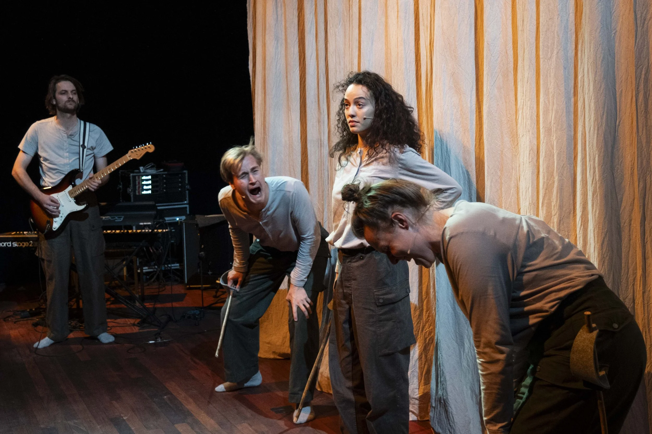 A theatrical performance with four women on stage in front of a yellow curtain. One woman stands with her hands on her hips, looking serious. The other three women are leaning forward with exaggerated expressions, one with her mouth open as if yellin