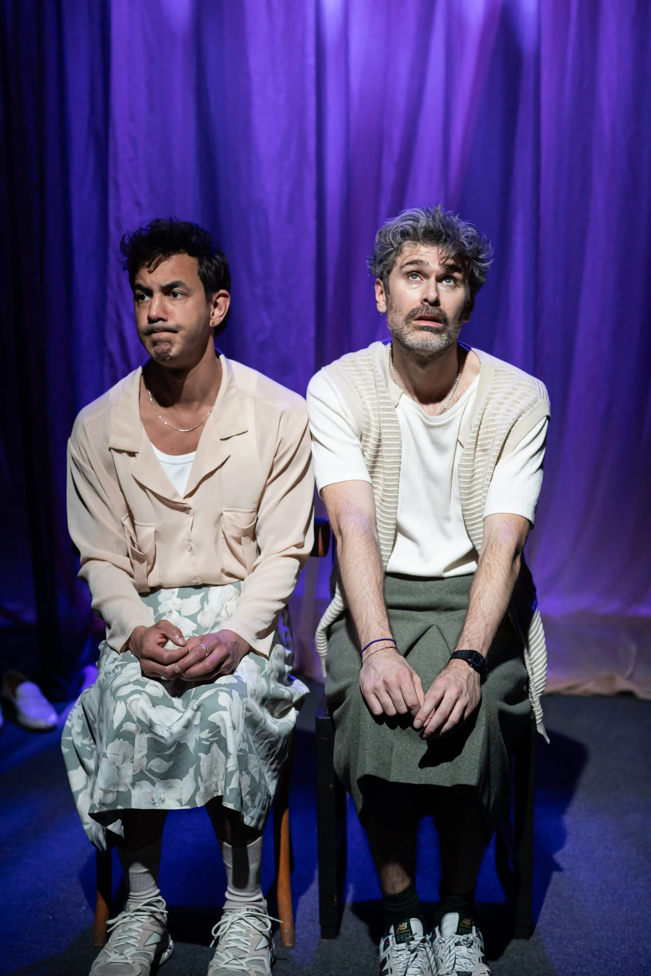 Two men sitting on chairs on a stage with purple curtains behind them, wearing casual clothing and looking contemplative.