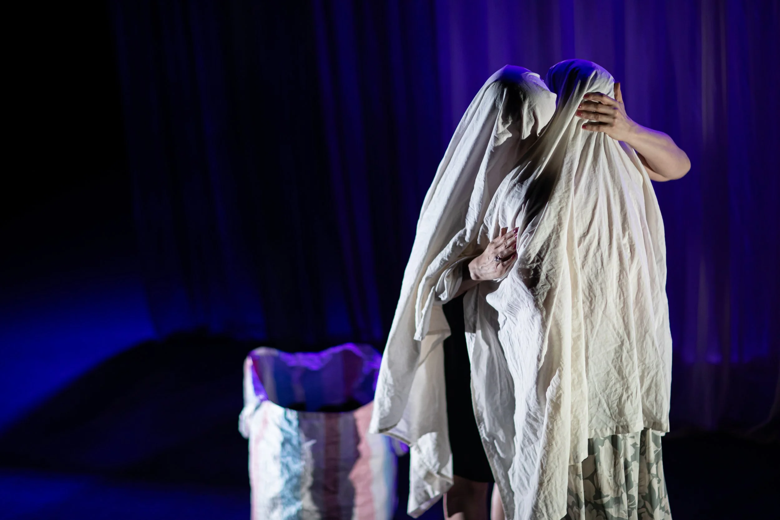 Two people on stage embrace, covered with white sheets, with a basket nearby, against a dark background with purple lighting.