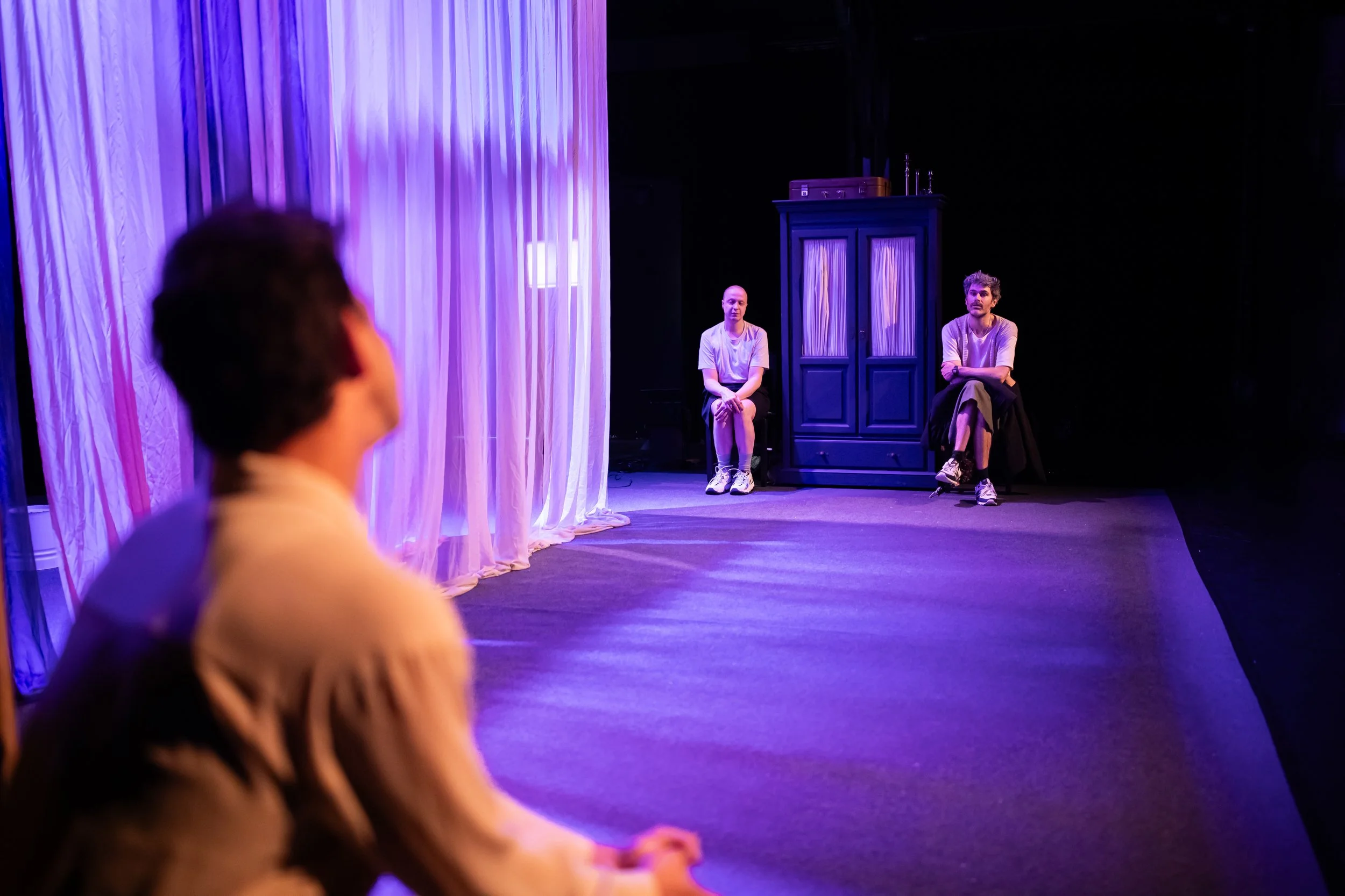 Three people on a stage with purple lighting and curtains, two sitting apart and one blurred in the foreground.