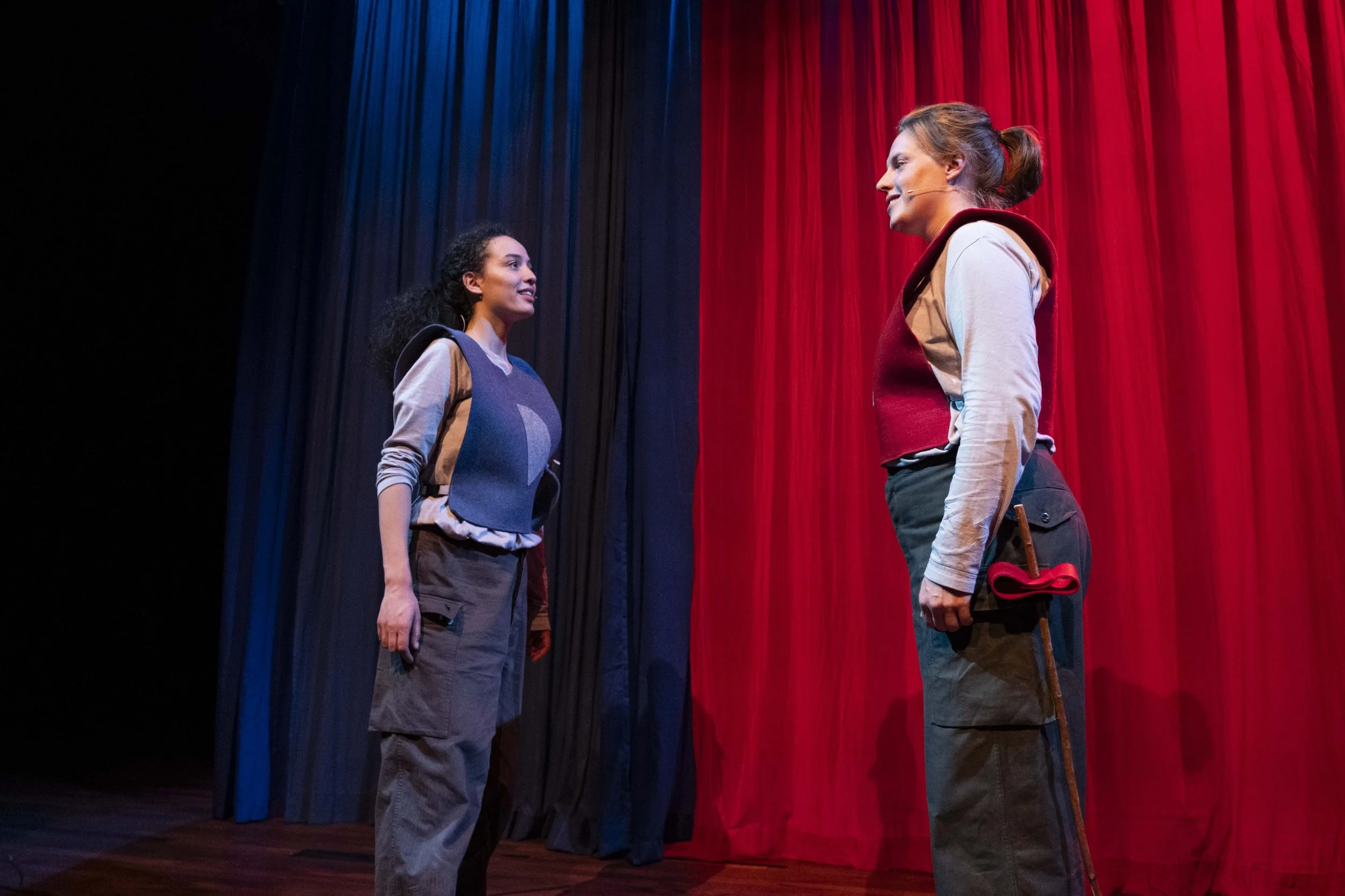 Two women on stage, facing each other, with a red and black curtain backdrop.