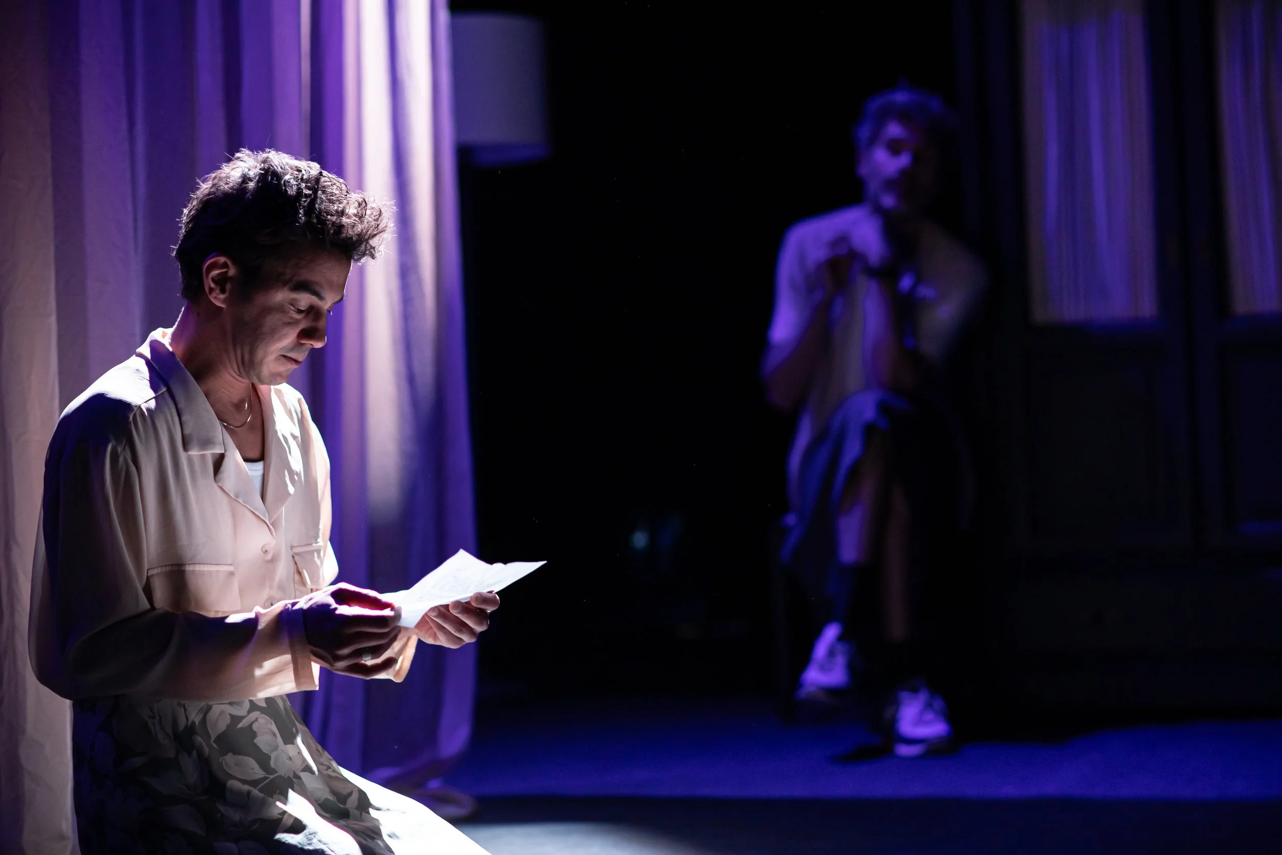 Two actors on stage, one in the foreground reading a paper and one in the background, sitting with knees up, both illuminated by stage lighting with curtains behind them.
